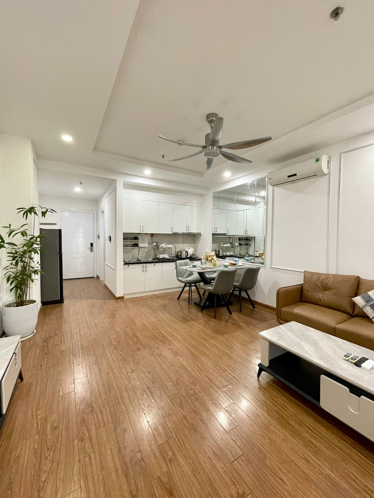 A modern living space features a brown sofa and a white coffee table. A dining area with a table and chairs is visible, alongside a fully equipped kitchen. Natural light brightens the room, enhanced by a ceiling fan and a potted plant.