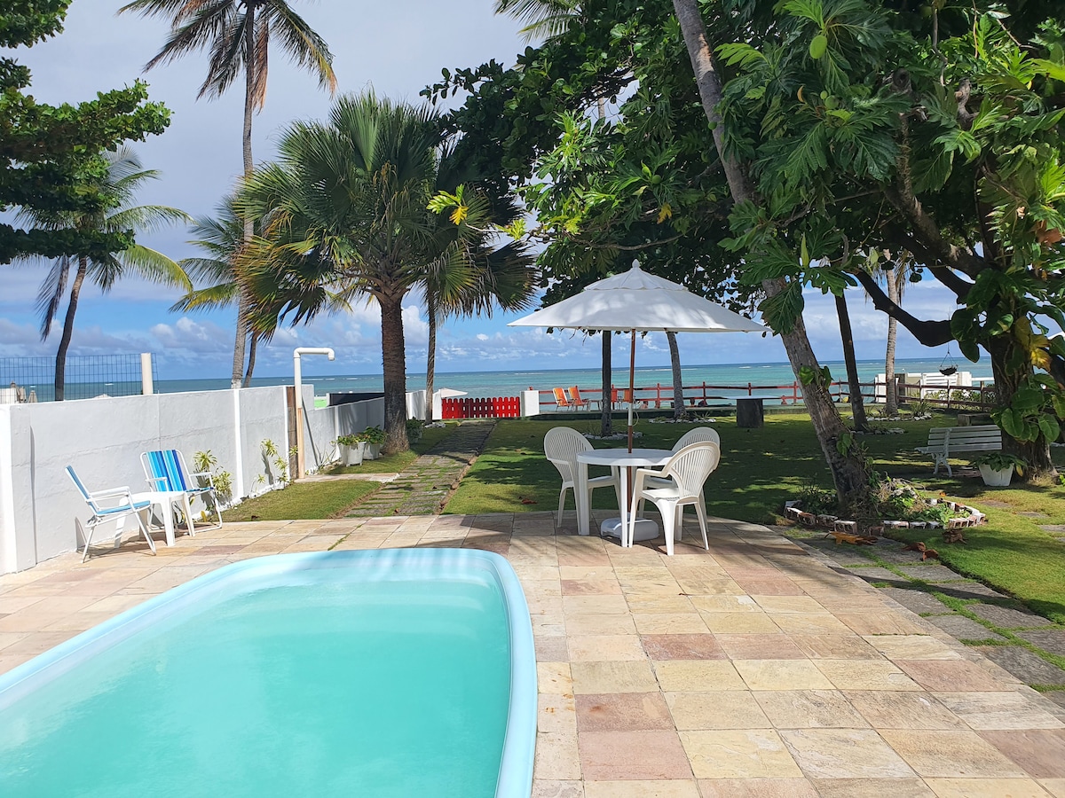A refreshing pool is set amid a spacious outdoor area, featuring a well-maintained lawn, trees, and beach access in the background. A shaded table with chairs sits nearby, while lounge chairs are positioned near the pool, providing ample space for relaxation.