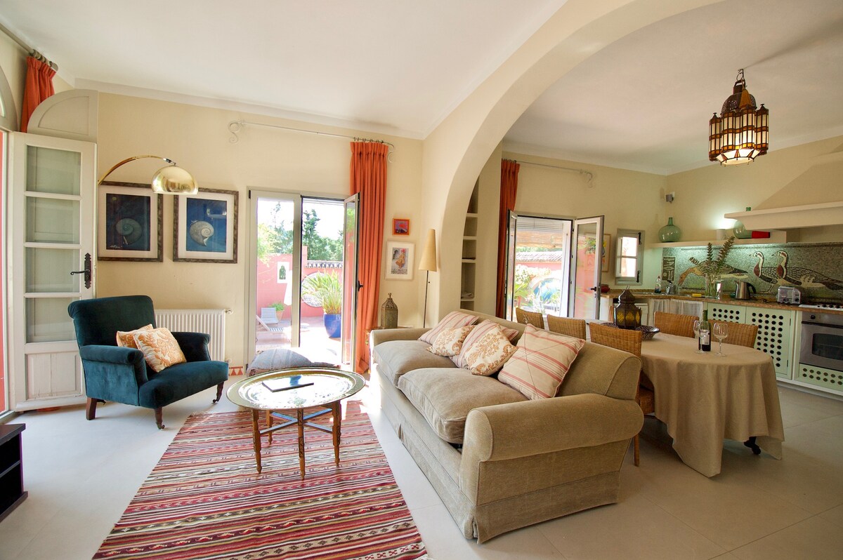 The open-plan living area features a comfortable sofa and a cozy armchair beside a circular table. Light floods in through multiple glass doors, leading to the outdoor area. Decorative elements include colorful cushions and artwork, creating a relaxed ambiance.