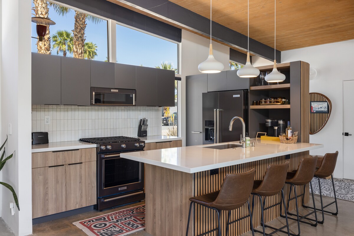 A modern kitchen showcases sleek cabinetry in dark tones and warm wood accents. A spacious island with seating is complemented by stylish pendant lighting. Updated appliances, including a gas stove and a built-in refrigerator, enhance the contemporary ambiance, while large windows offer views of the outdoors.