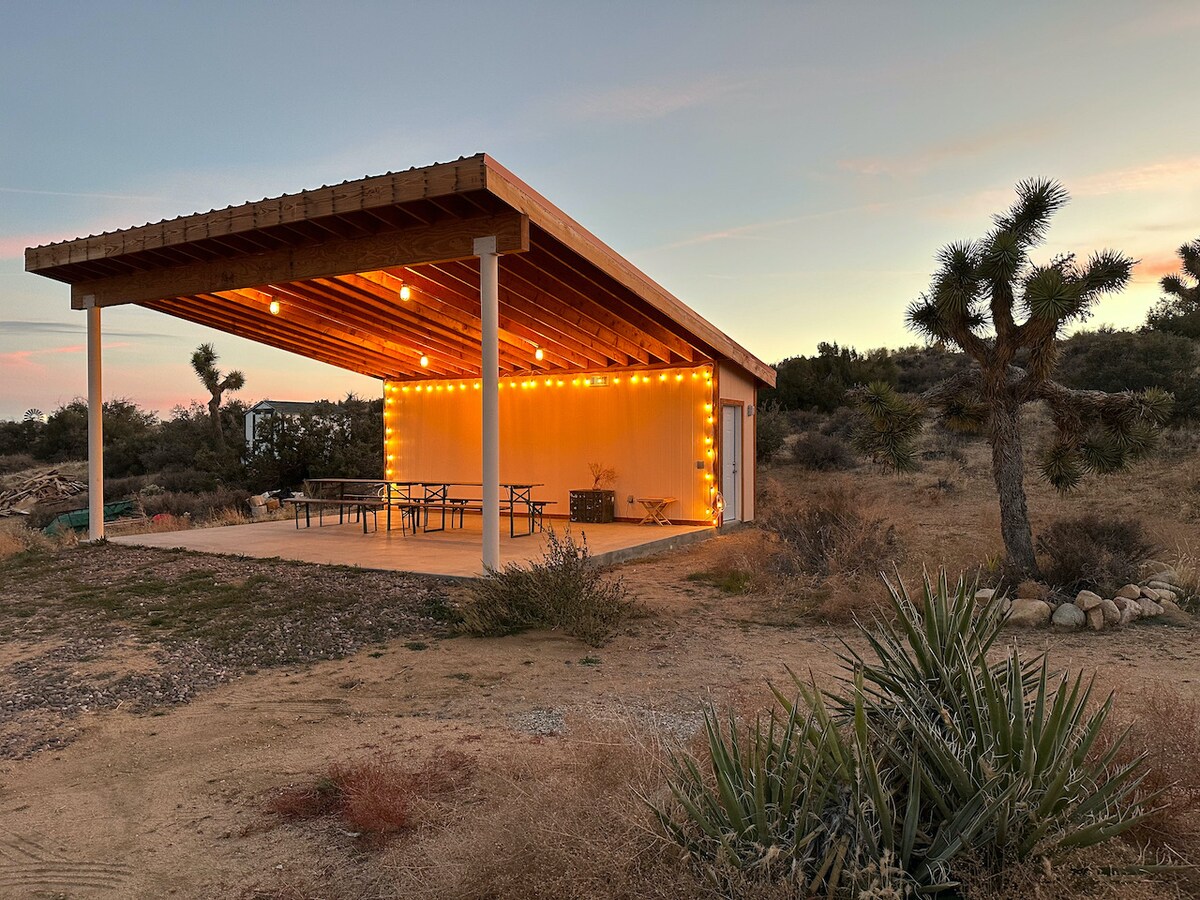 An outdoor patio area features a wooden roof supported by columns, illuminated by string lights. Large tables and seating are provided under the shelter, surrounded by desert vegetation. The sunset casts a warm glow, enhancing the tranquil setting.