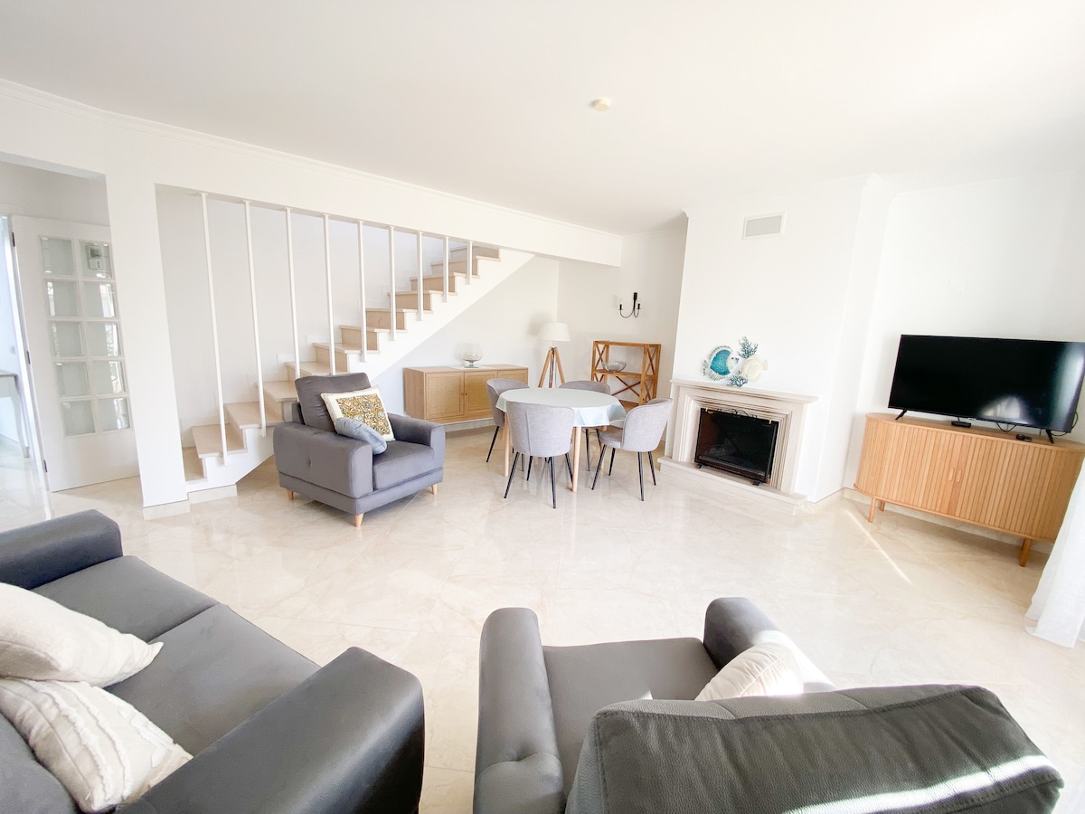 A spacious living area features comfortable seating arrangements with soft gray sofas and stylish armchairs. A round dining table is surrounded by elegant chairs. Natural light is allowed through large windows, highlighting the light-colored flooring and minimalist decor, including a television and decorative elements.