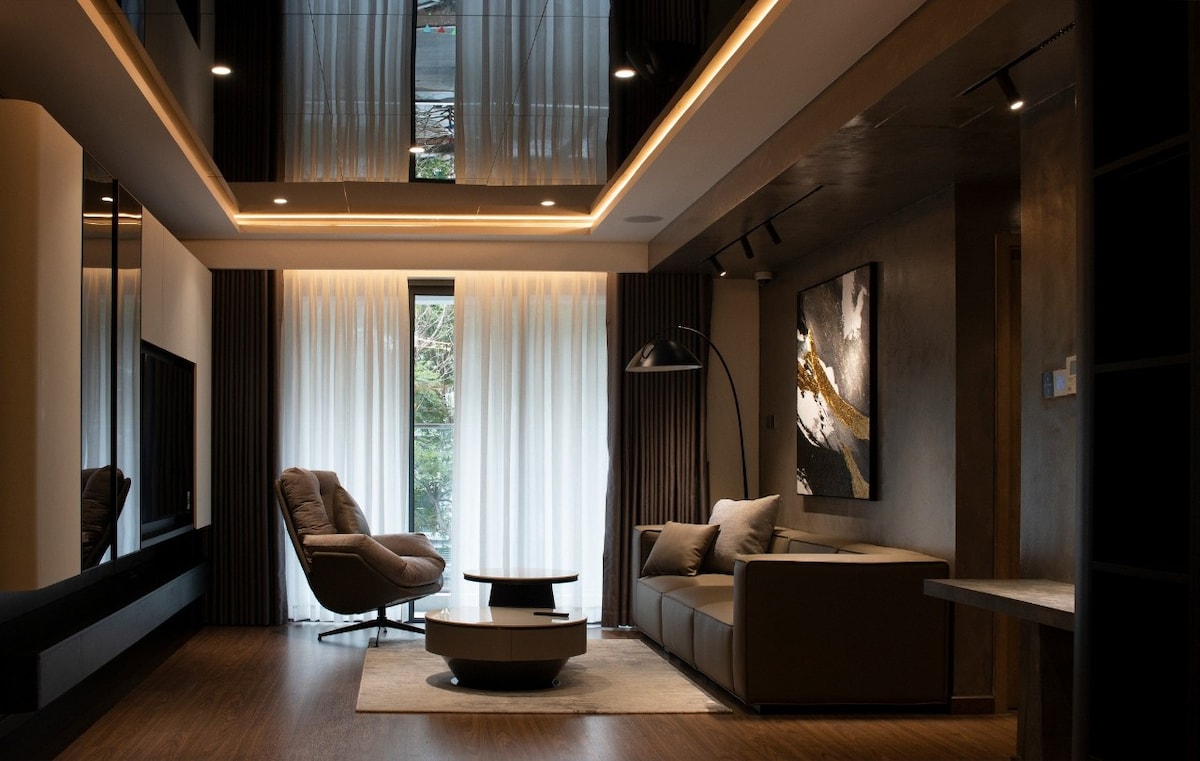 A modern living area features large windows adorned with sheer curtains, allowing natural light to fill the space. A comfortable sofa is positioned next to a round coffee table, and an armchair is placed nearby, creating inviting seating arrangements. Light fixtures enhance the contemporary decor.