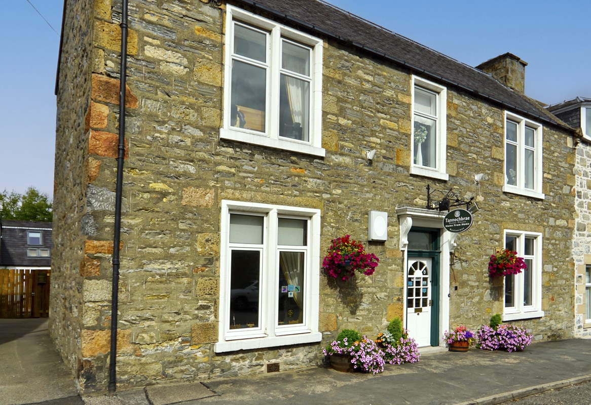 The exterior of a charming stone house features a classic design with large windows adorned with flower boxes. Vibrant flowers add color along the entrance, while the plaque indicates the property's name. The structure is set against a clear blue sky.
