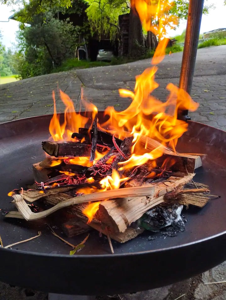 A fire burns brightly in a metal fire bowl, with flames reaching upwards and illuminating the surrounding area. Stacked firewood forms a base, creating a warm atmosphere for shared evenings outdoors.