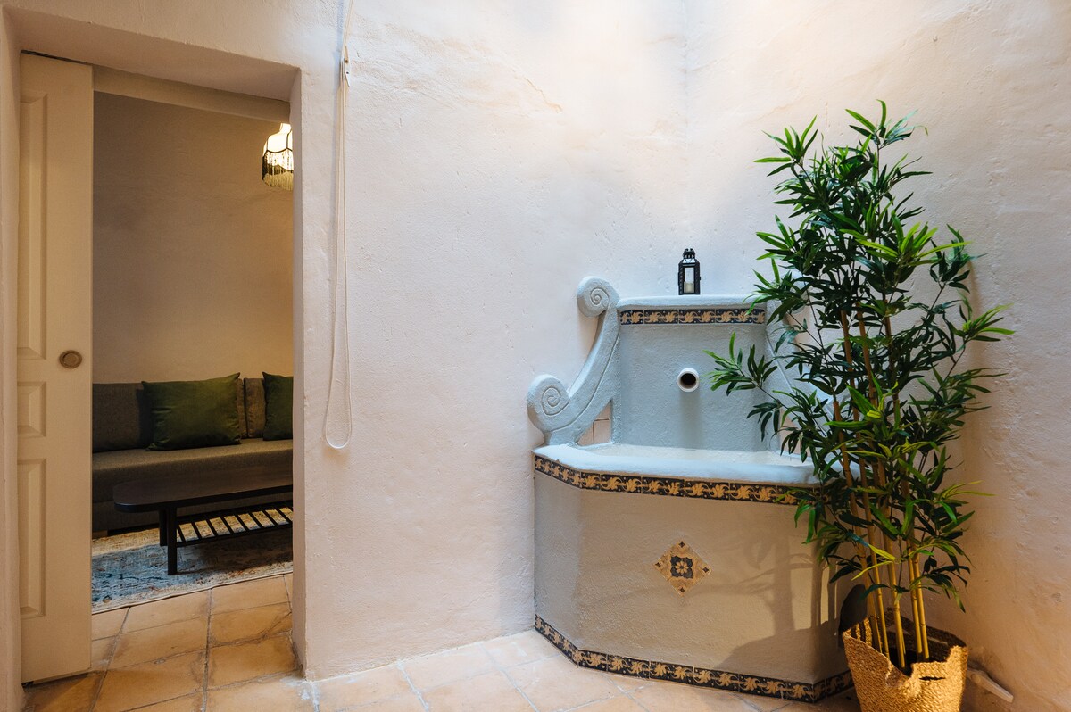 An interior space showcases a decorative stone fountain against a textured white wall. A potted plant adds a touch of greenery beside the fountain. A cozy seating area is visible in the background, featuring a modern sofa with plush cushions.