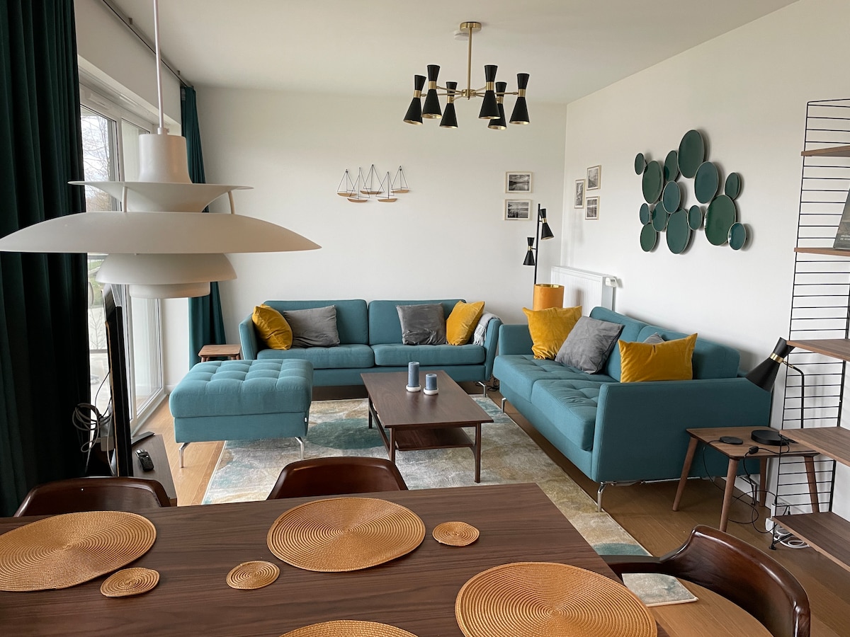 A spacious living area is showcased, featuring a large turquoise sectional sofa paired with cushions. An elegant dining table is set with round placemats, while modern light fixtures hang above. Art pieces and decorative elements are arranged on the wall, providing visual interest.