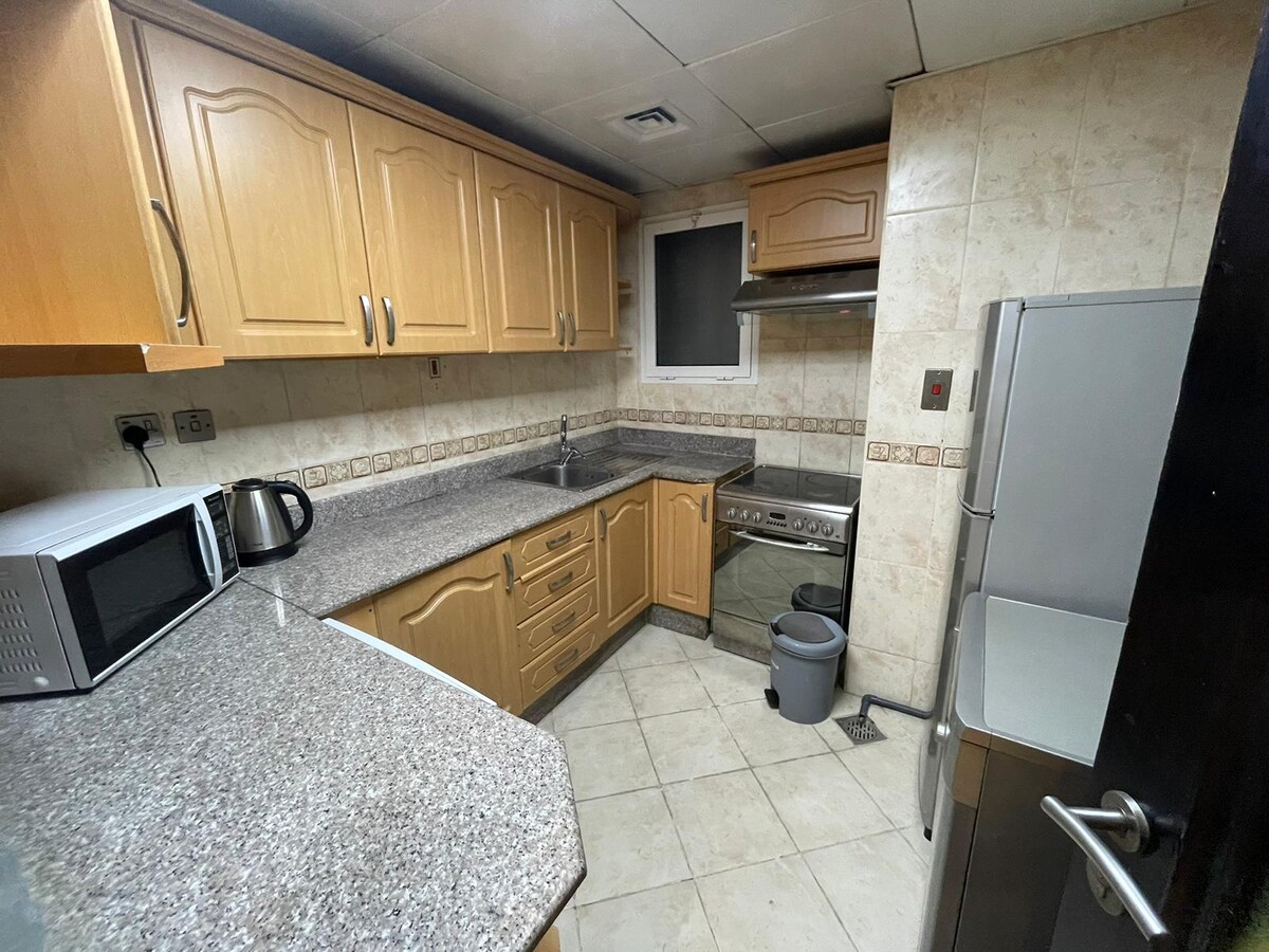 The kitchen is equipped with wooden cabinetry and granite countertops. Appliances include a microwave, kettle, and stove. A stainless steel refrigerator is present, along with a small garbage bin. Natural light enters through a window, illuminating the tiled floor.
