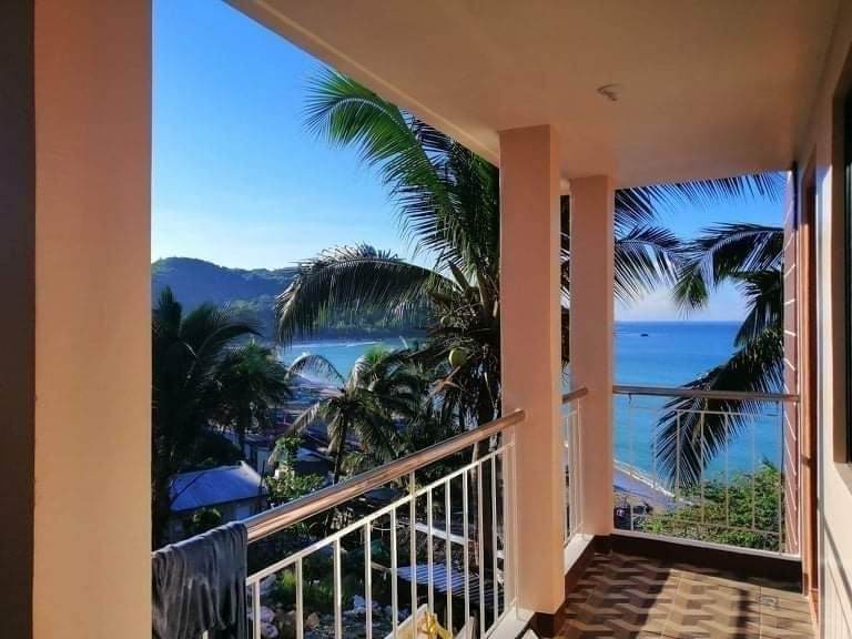 A balcony view is presented with palm trees gently swaying in the breeze, overlooking a serene seascape. The crystal-clear water meets the shoreline, while distant hills create a tranquil backdrop.