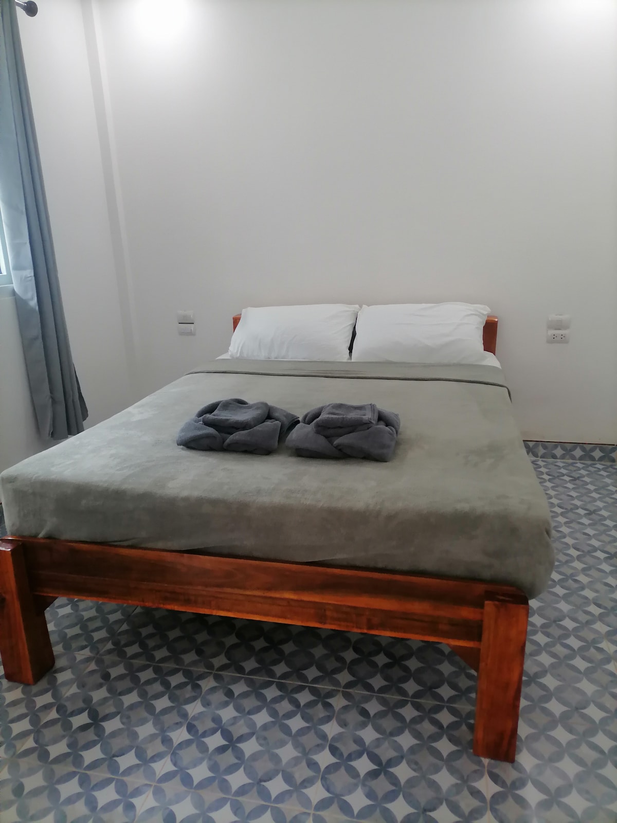 A simple sleeping area features a spacious bed with light-colored linens atop a wooden frame. Two neatly folded towels are placed on the bed, and patterned tiles cover the floor. Natural light enters through a nearby window, enhancing the serene ambiance.