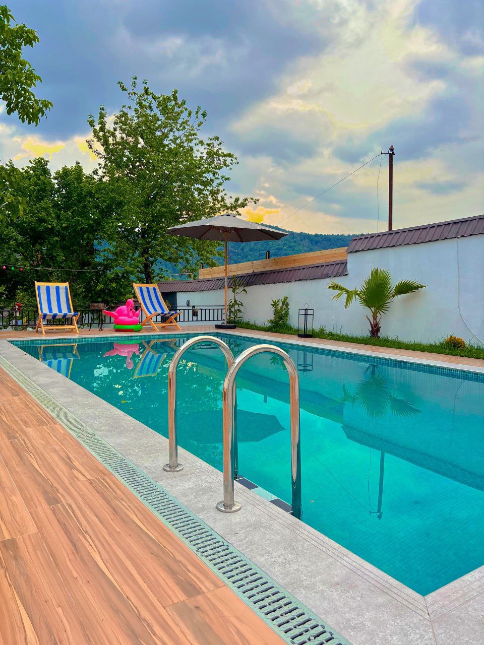 A private pool is surrounded by lush greenery and features two lounging chairs under an umbrella. The clear water of the pool reflects the sky, and a pink float adds a pop of color. A section of the patio is lined with wooden decking.