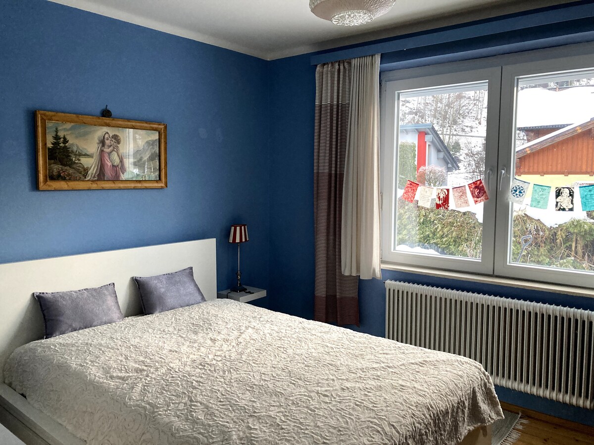 A serene bedroom is presented with blue walls and a spacious bed adorned with a light quilt. A framed painting hangs above the bed, while natural light enters through a large window dressed with sheer curtains. Decorative items can be seen strung along the window.