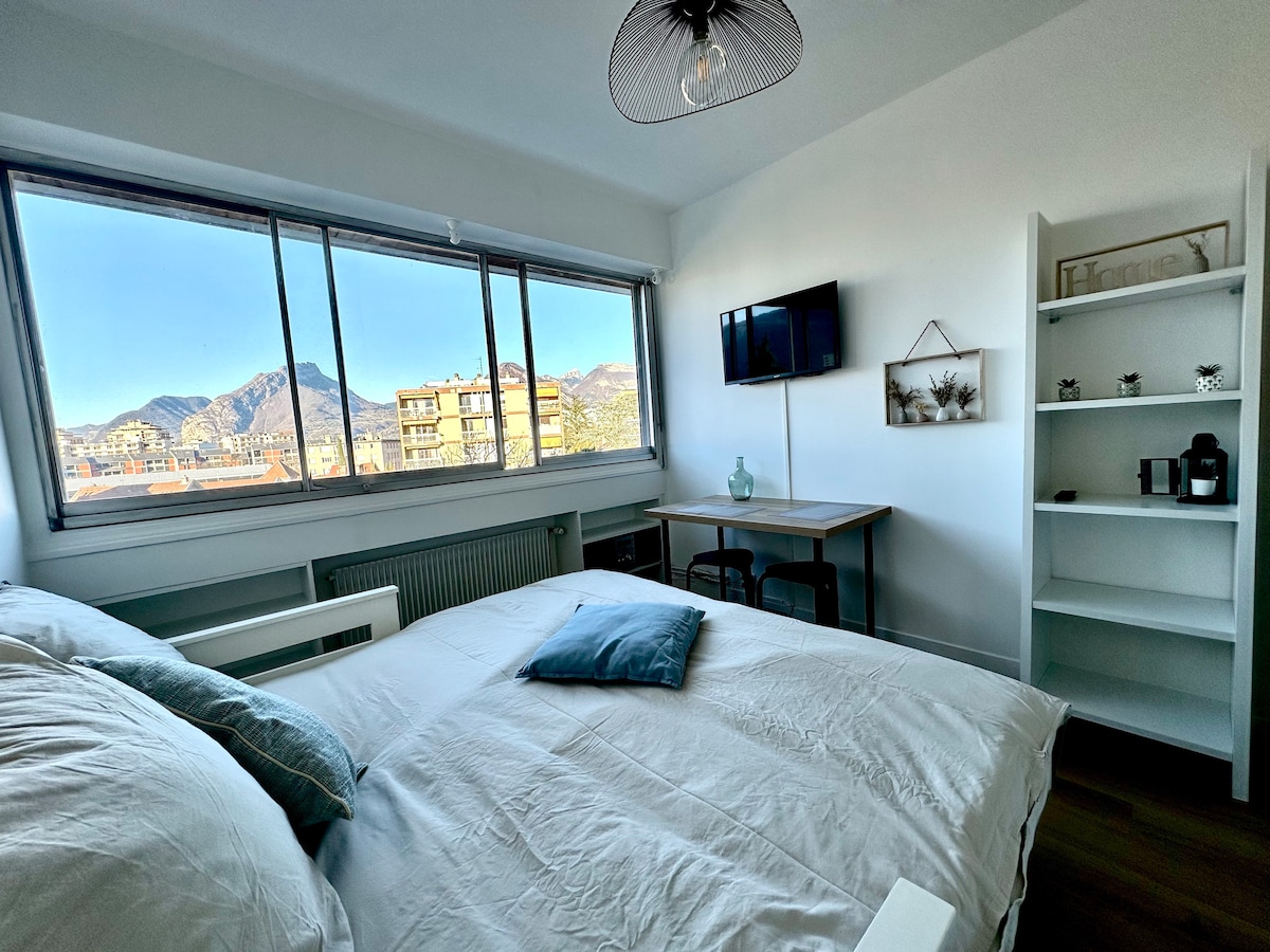 A bright sleeping area is presented, featuring a spacious bed with soft linens and decorative pillows. Large windows offer a panoramic view of the mountainous landscape. A wall-mounted TV is visible, along with a small desk and a shelf containing decorative items and a kettle.