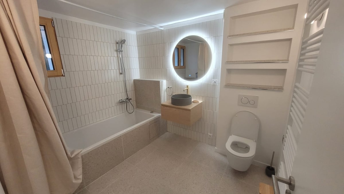 A modern bathroom features neutral-colored tiles and a spacious bathtub paired with a handheld shower. A circular mirror with built-in lighting is mounted above a minimalist vanity. Natural light is allowed in through a nearby window, with a privacy curtain partially drawn.