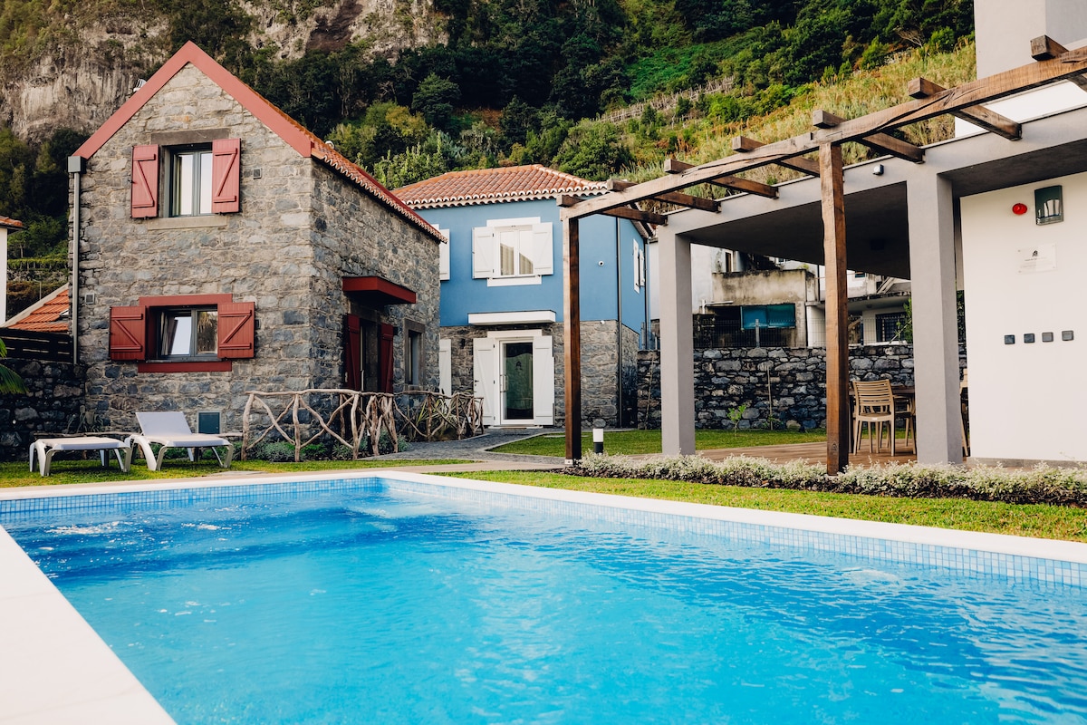 A tranquil outdoor area is showcased, featuring a clear blue swimming pool bordered by neatly maintained grass. Two independent stone houses with colorful shutters are visible, along with a covered dining area under a modern pergola. Lush greenery and mountains are seen in the background.