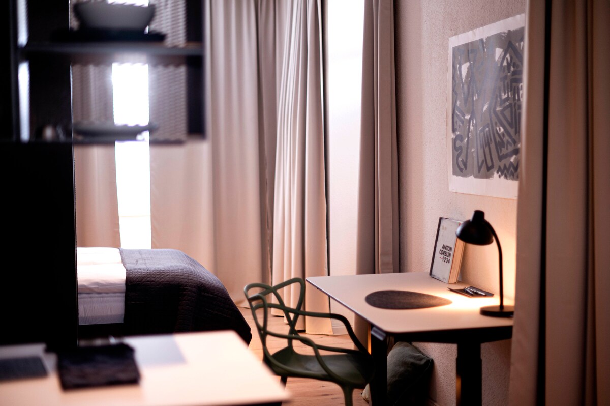 An open living space features a spacious bed with neutral bedding, a modern work desk with a black lamp, and a colorful chair. Light filters through sheer curtains, highlighting the contemporary decor and inviting ambiance.