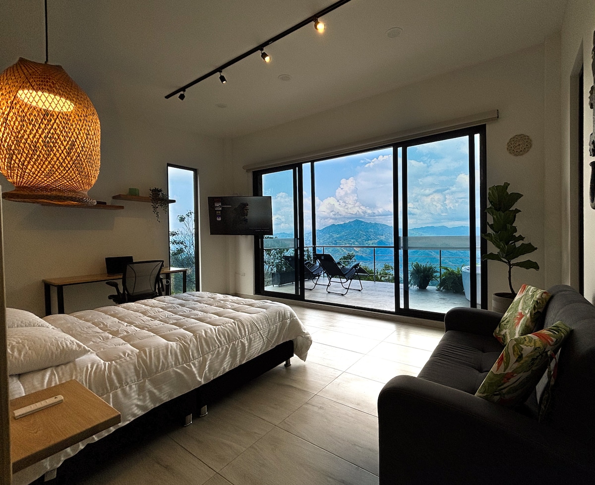 A spacious room features large glass doors opening to scenic mountain views. A comfortable queen bed and a sofa are positioned within the area, while a small workspace with a desk is visible. Natural lighting enhances the serene atmosphere.