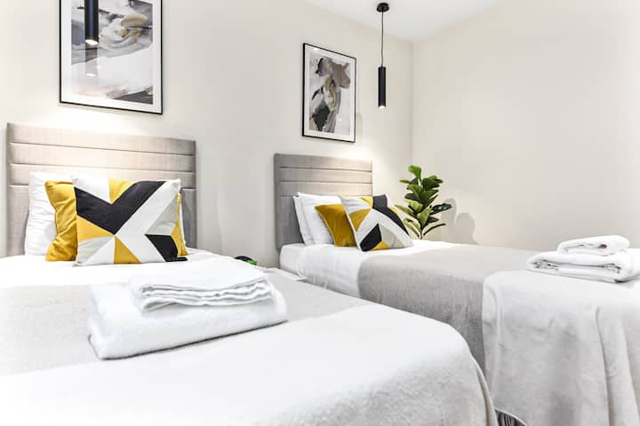 Disfruta de la elegancia de la ciudad en este elegante dormitorio con dos cómodas camas individuales, un elegante escritorio y un televisor montado en la pared perfectamente posicionado para trabajar y relajarse.