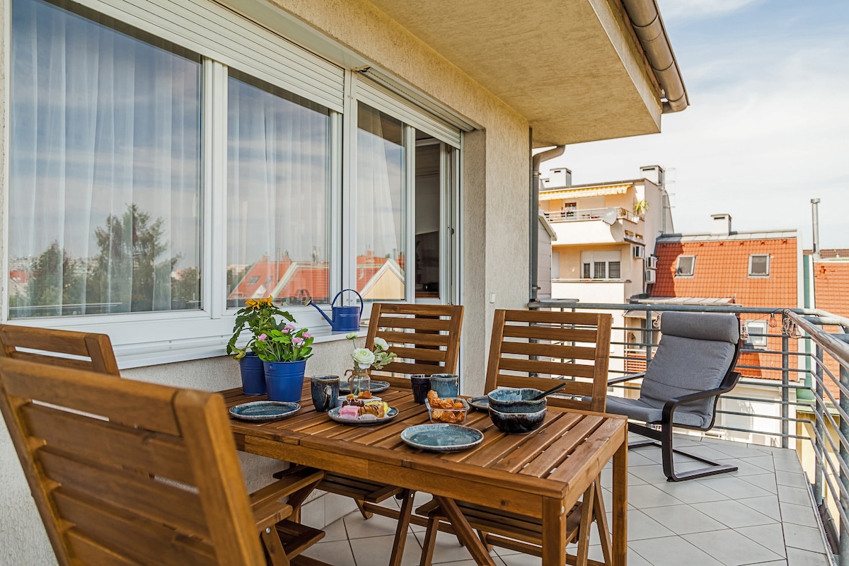 A charming outdoor balcony features a wooden dining table surrounded by comfortable chairs. Potted plants and decorative items add a personal touch. The view includes rooftops and distant buildings under a clear sky, creating a serene outdoor dining space.