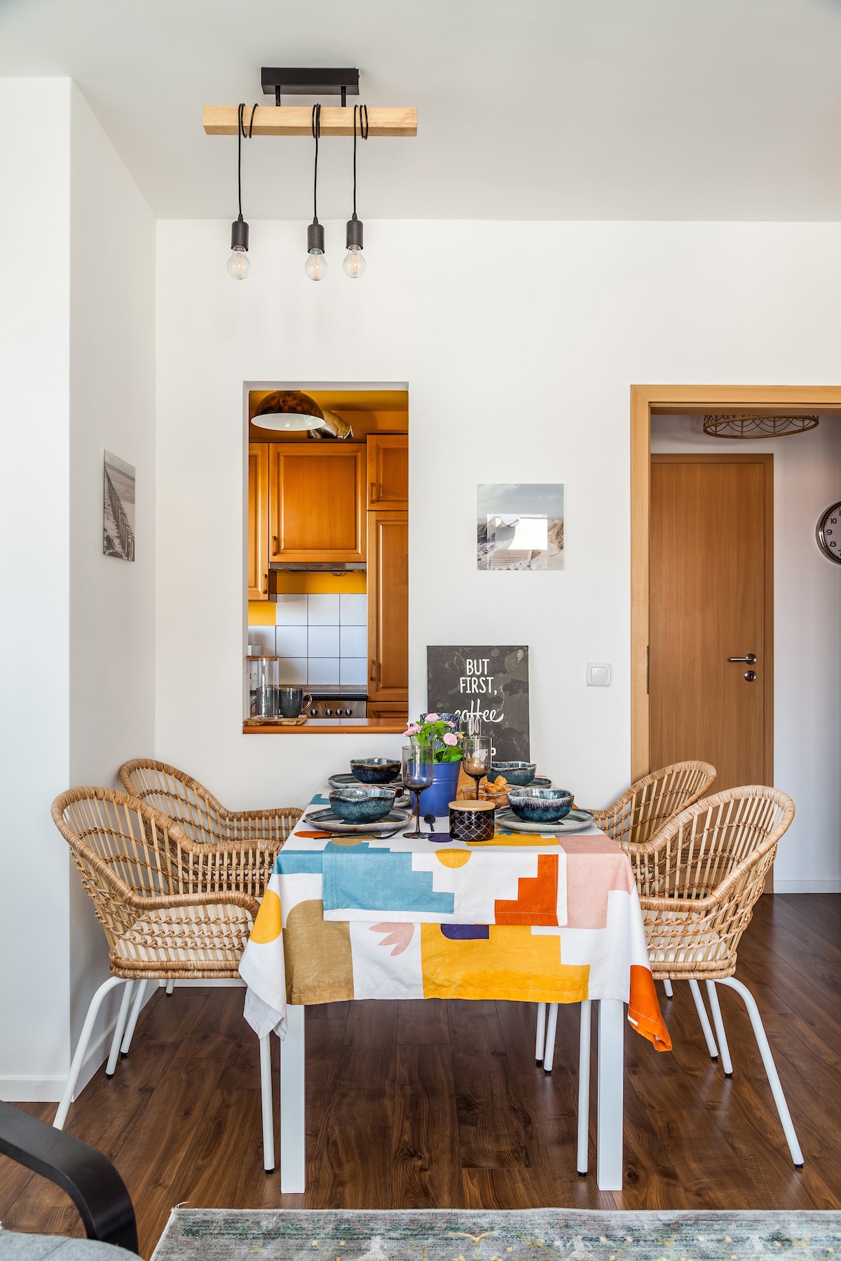 A dining area featuring a colorful tablecloth covers a table set for a meal. Four wicker chairs surround the table, which is adorned with tableware and a small vase of flowers. A warm wood kitchen can be seen through a serving window.