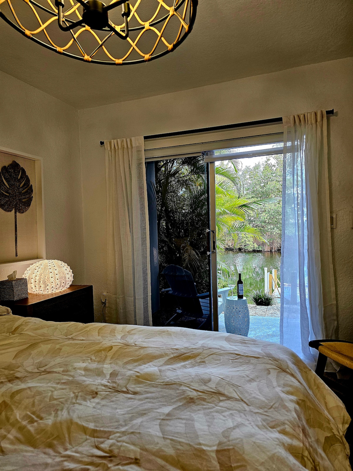A serene bedroom features a large sliding glass door leading to a tranquil outdoor space with water and greenery visible beyond. A bedside lamp provides soft lighting, and a spacious bed is dressed in light-colored bedding. Drapes hang gently by the door.