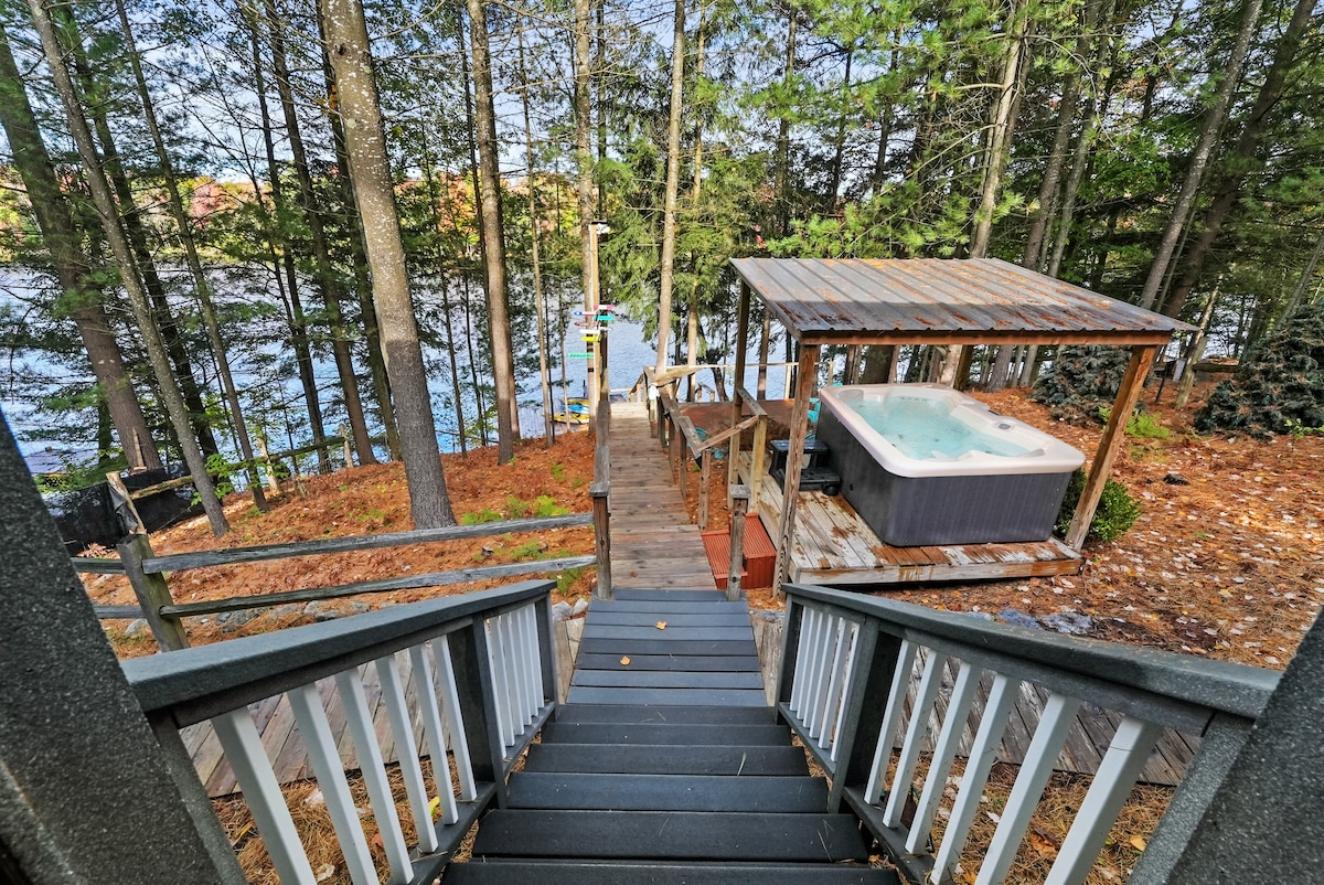 Wooden steps lead down through a canopy of pine trees towards the lake. To the right, a hot tub is sheltered under a wooden gazebo, while further ahead, a large dock extends into the calm water.