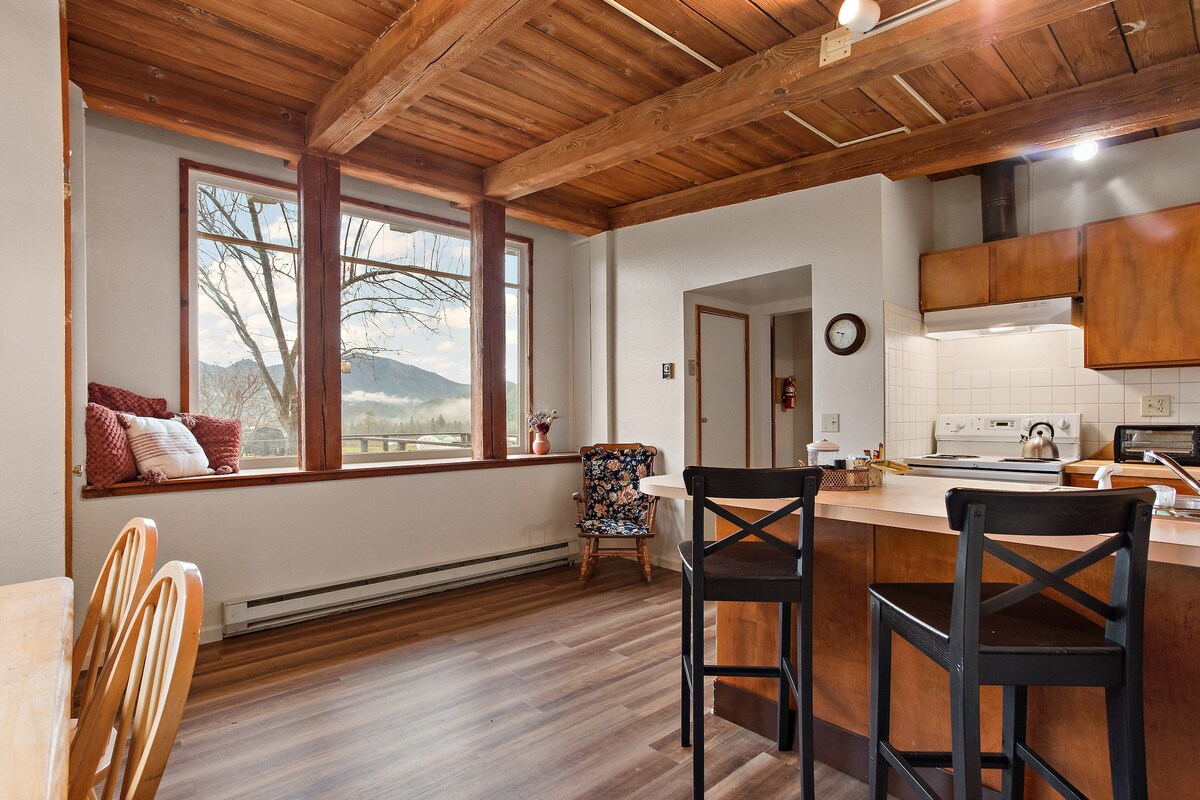 A welcoming kitchen area features wooden beams and large windows that provide views of the surrounding landscape. A circular dining table and two chairs are positioned at one side, while the kitchen counter includes bar stools for additional seating. A cozy nook with cushions is visible near the windows.