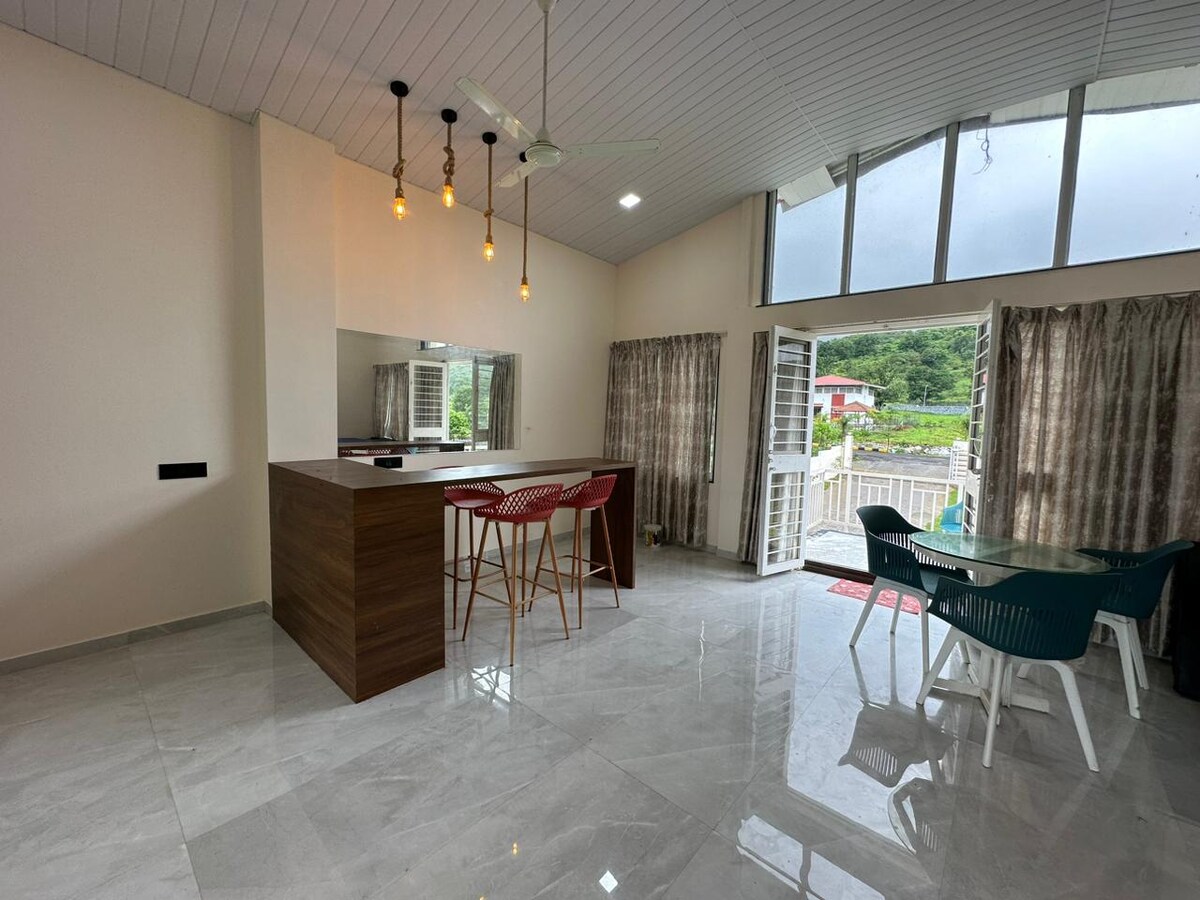 An open-plan living area is visible, featuring a modern kitchen with a wooden island and high chairs. Large windows allow natural light to fill the space, while sliding doors lead to an outdoor seating area. Two sets of curtains frame the windows, enhancing the room's ambiance.