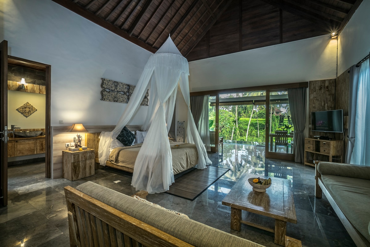 A spacious villa interior features a king-sized bed draped with a white canopy. A comfortable seating area with wooden furniture is present. Large glass doors provide a view of the surrounding greenery, while a small kitchenette is visible in the background.