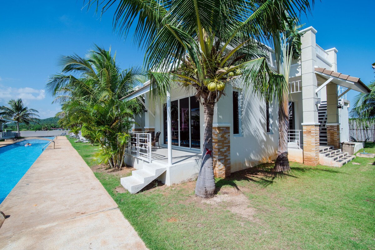 A modern villa is viewed from the side, featuring a poolside patio with a white railing. Lush palm trees provide shade around the outdoor space. A turquoise swimming pool extends alongside the villa, bordered by a neatly maintained lawn and a paved walkway.