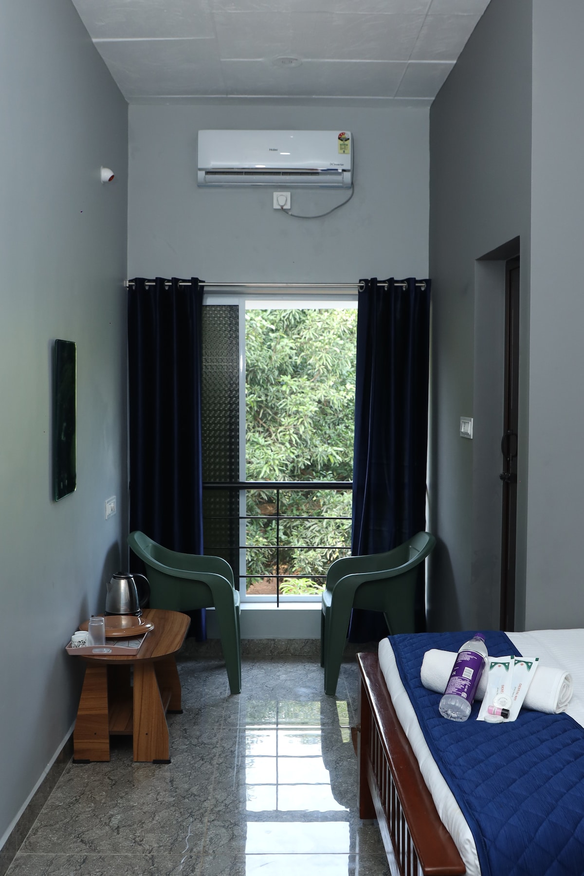 A cozy corner of the room features a spacious bed beside a wooden table and two green chairs. Large windows provide a view of the garden, with dark curtains enhancing privacy. An air conditioning unit is mounted above, ensuring a comfortable environment.