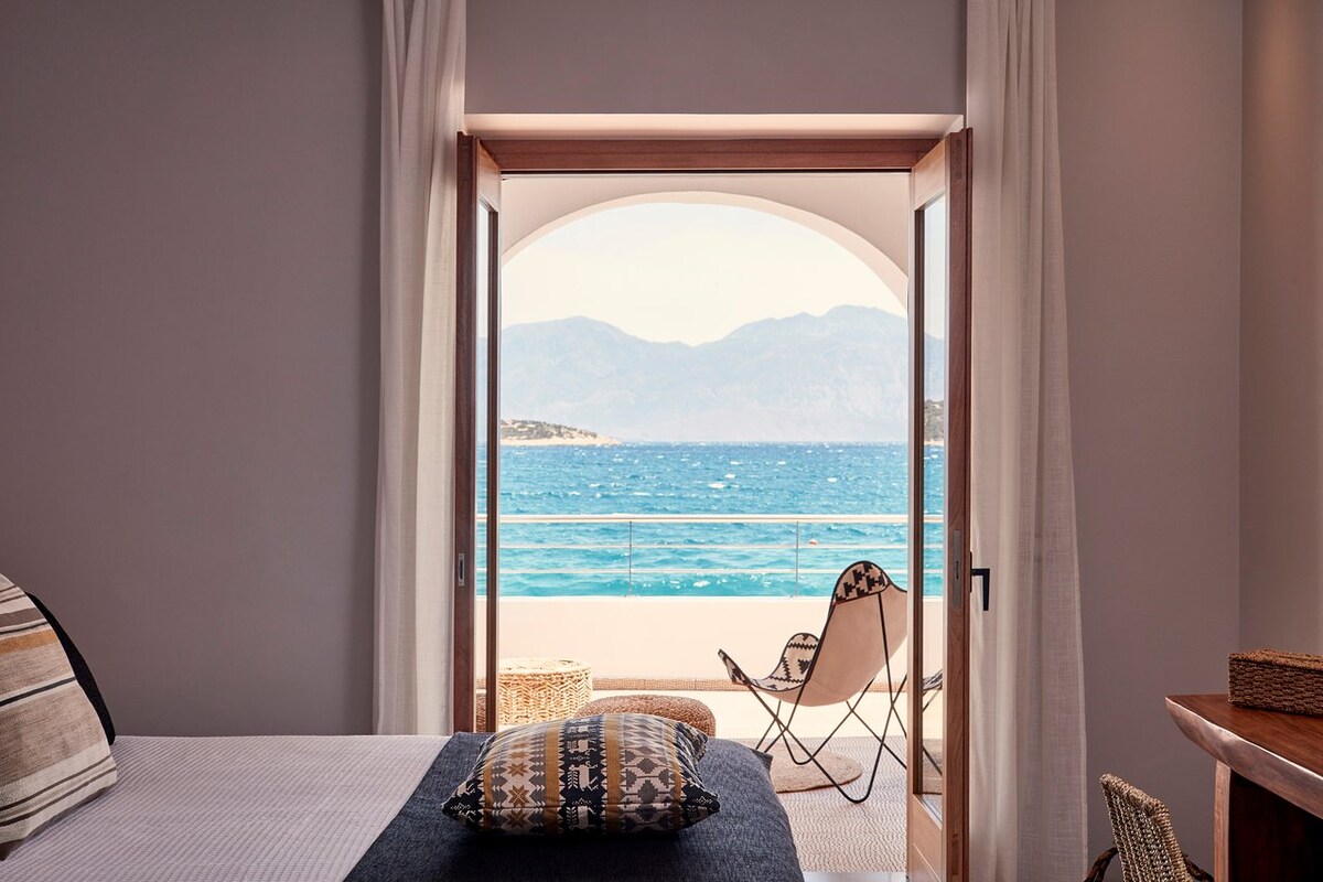 A spacious room features an open doorway revealing a terrace overlooking the sea. Bright sunlight illuminates the interior, highlighting a textured bedspread and decorative pillows. A comfortable chair is positioned on the terrace, providing a spot to enjoy the scenic coastal view.