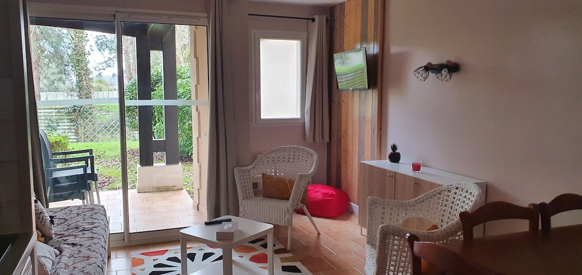 A bright living space is shown, featuring a comfortable seating area with two woven chairs and a small coffee table. Sliding glass doors lead to an outdoor terrace with garden views. A TV is mounted on the wall, complemented by wooden accents and natural light.