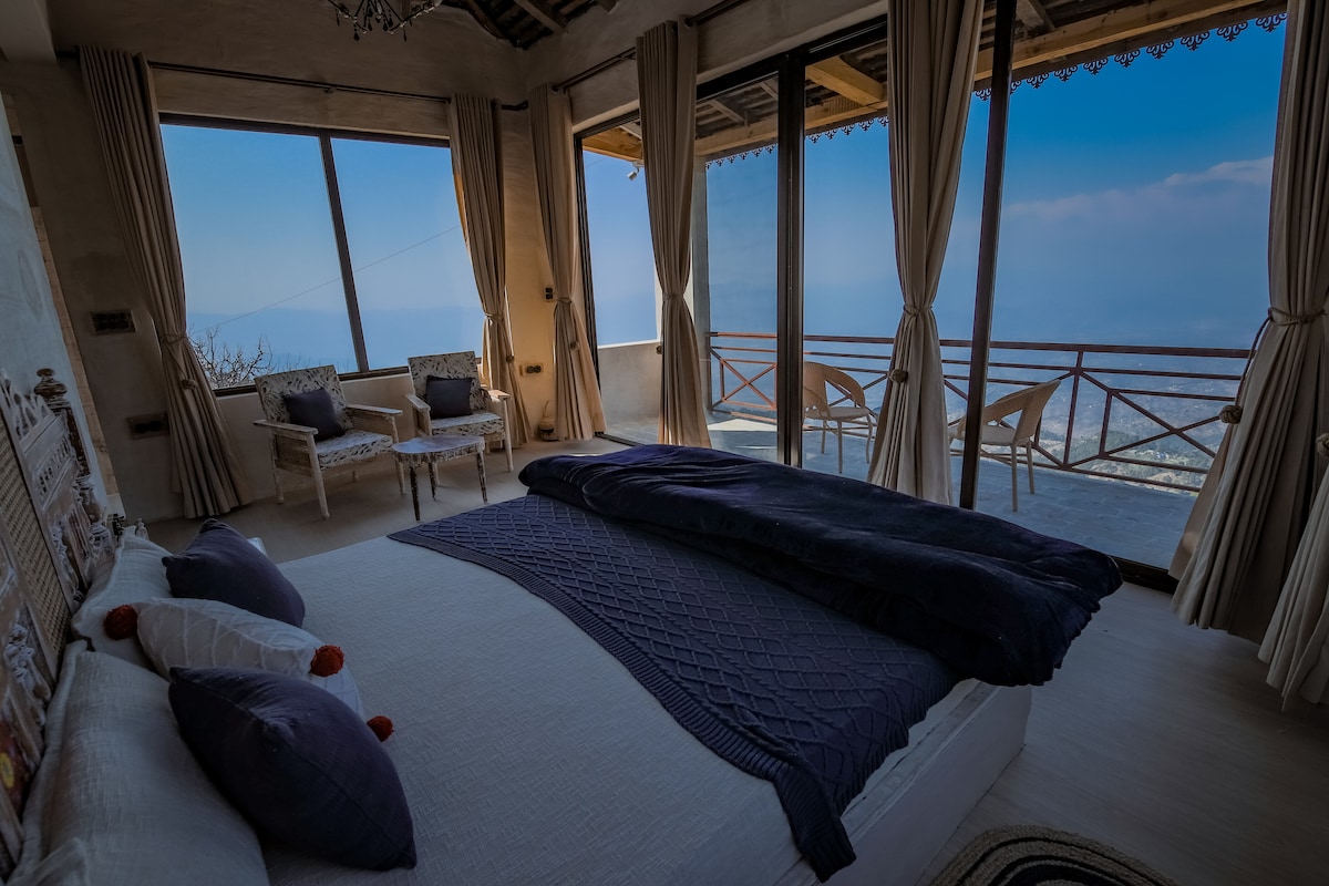 A spacious bedroom is highlighted by large windows offering panoramic views of the surrounding hills. A neatly made bed with a textured blanket is positioned centrally, while light-colored curtains frame the windows. Two chairs and a small table on the balcony enhance the serene atmosphere.