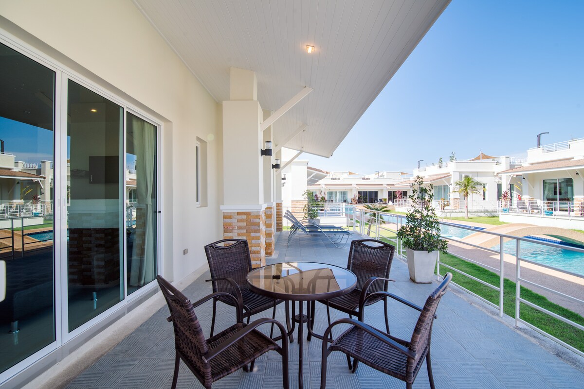 A shaded outdoor patio area is furnished with a round table and four wicker chairs, providing a comfortable space to relax. The patio opens to a well-maintained lawn and a glimpse of the sparkling pool beyond, framed by the villa's modern architectural lines.