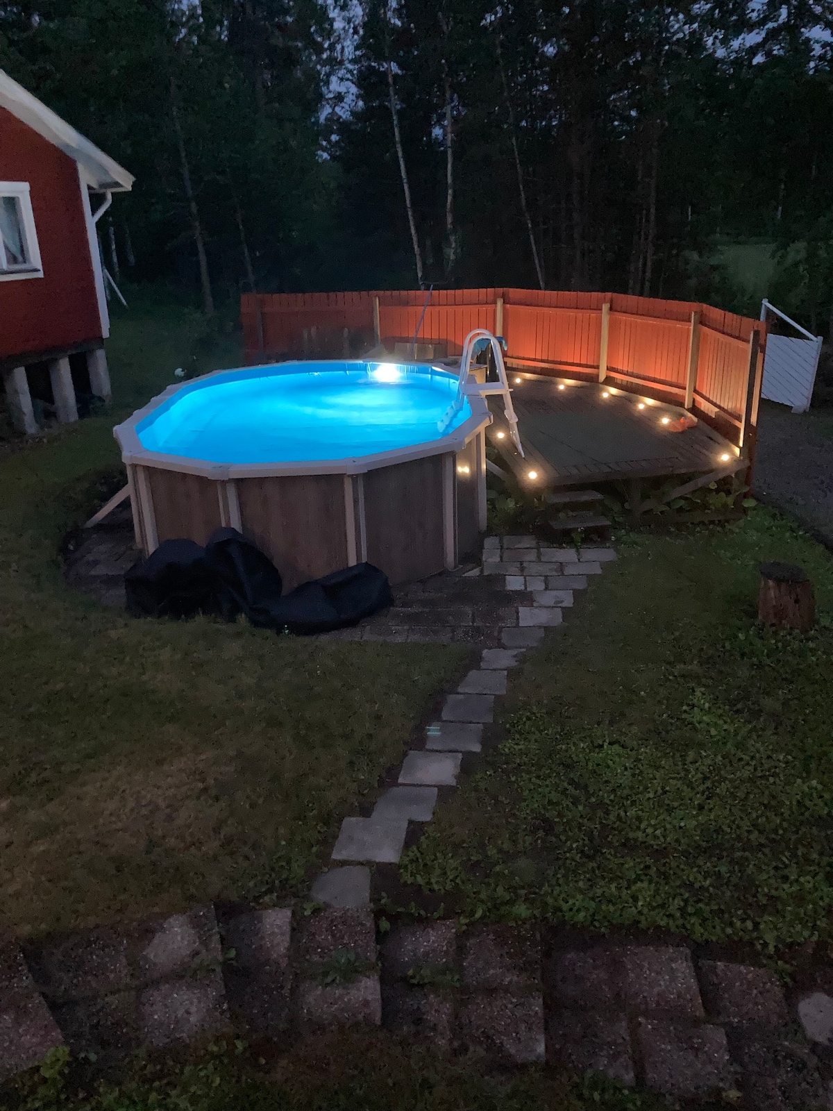 A heated above-ground pool is situated on a wooden deck, surrounded by softly glowing lights. The pool is illuminated in blue, creating a serene ambiance. A pathway of stones leads from the pool area, framed by lush grass and greenery.