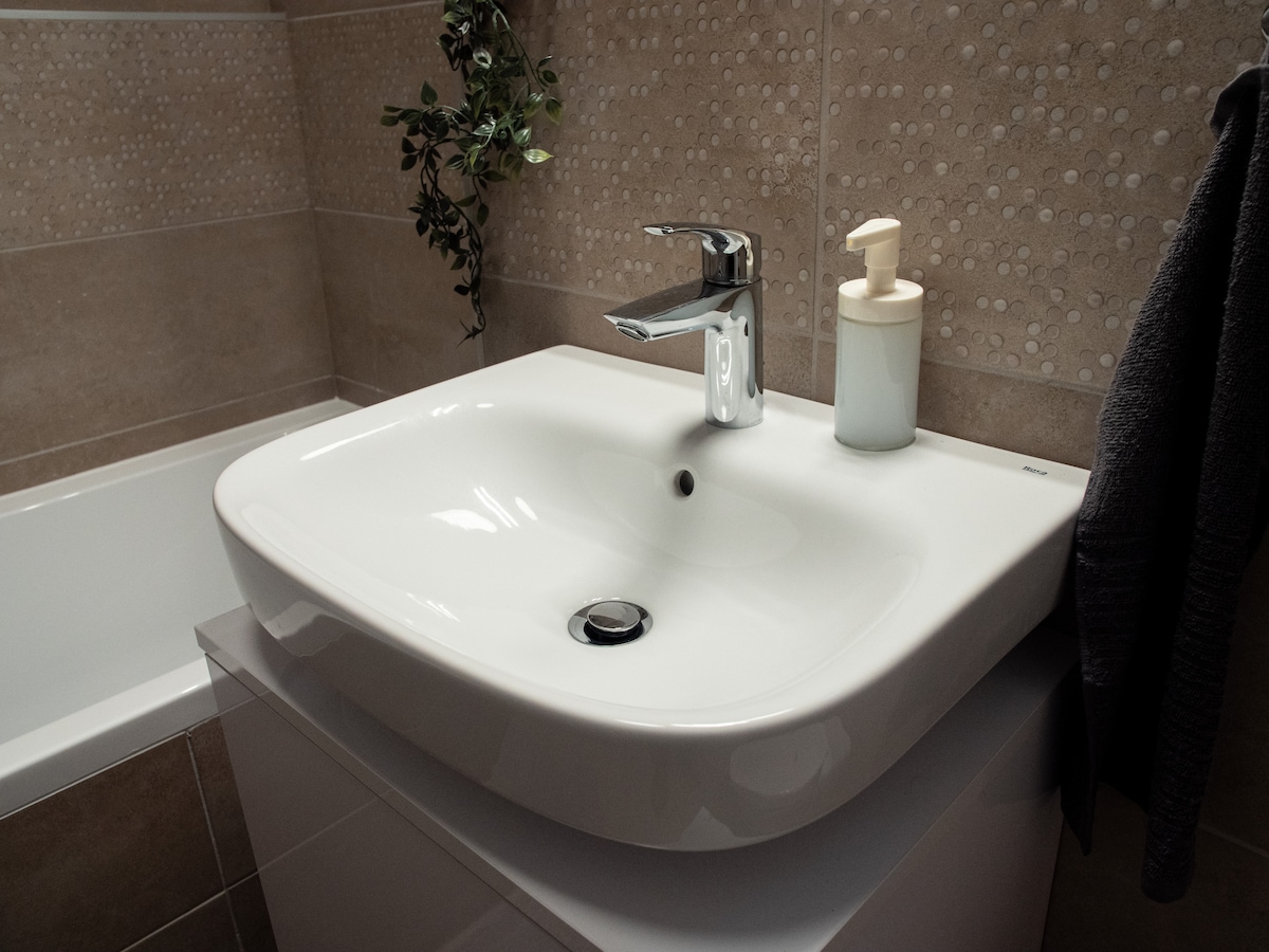 A modern bathroom sink is featured, showcasing a sleek faucet and a soap dispenser beside it. The sink is placed atop a white vanity, with a soft towel hanging nearby. The surrounding walls are elegantly tiled in neutral tones, contributing to a clean aesthetic.