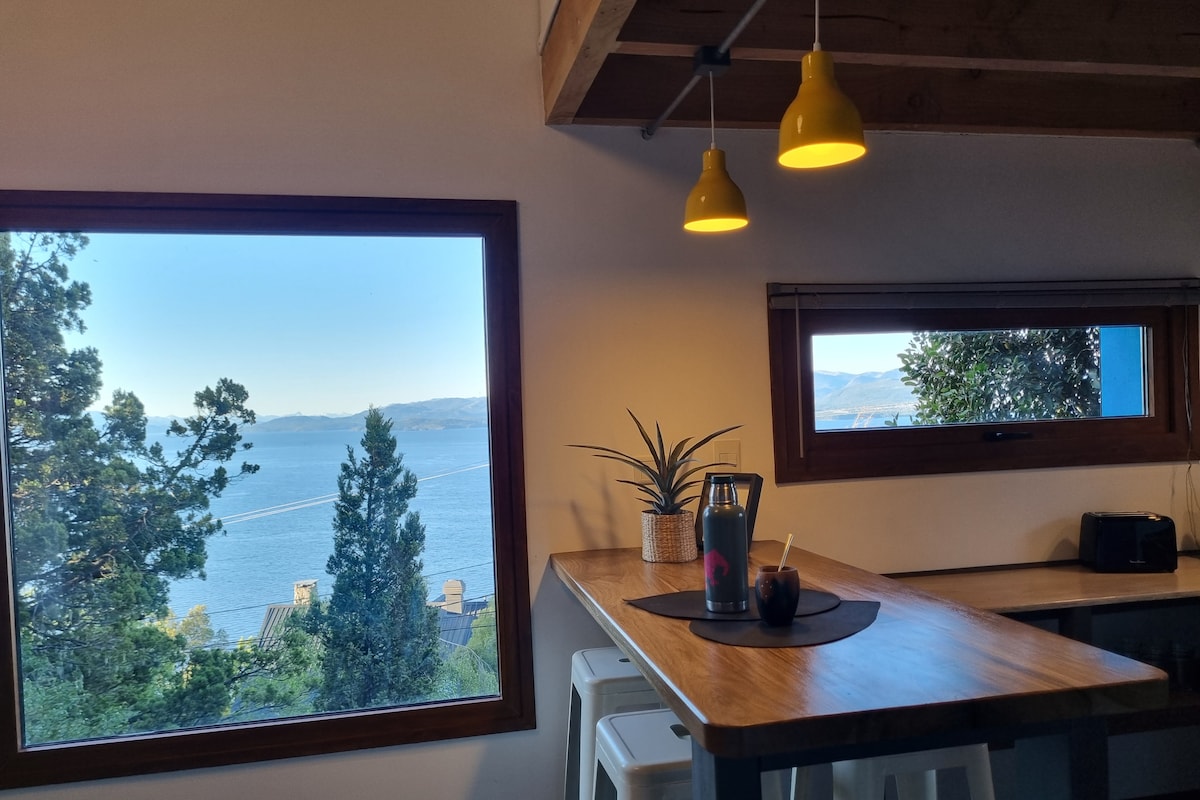 A dining area features a wooden table illuminated by yellow pendant lights. Large windows offer views of the lake and surrounding trees, enhancing the serene ambiance. A small plant and a beverage sit on the table, contributing to the cozy environment.