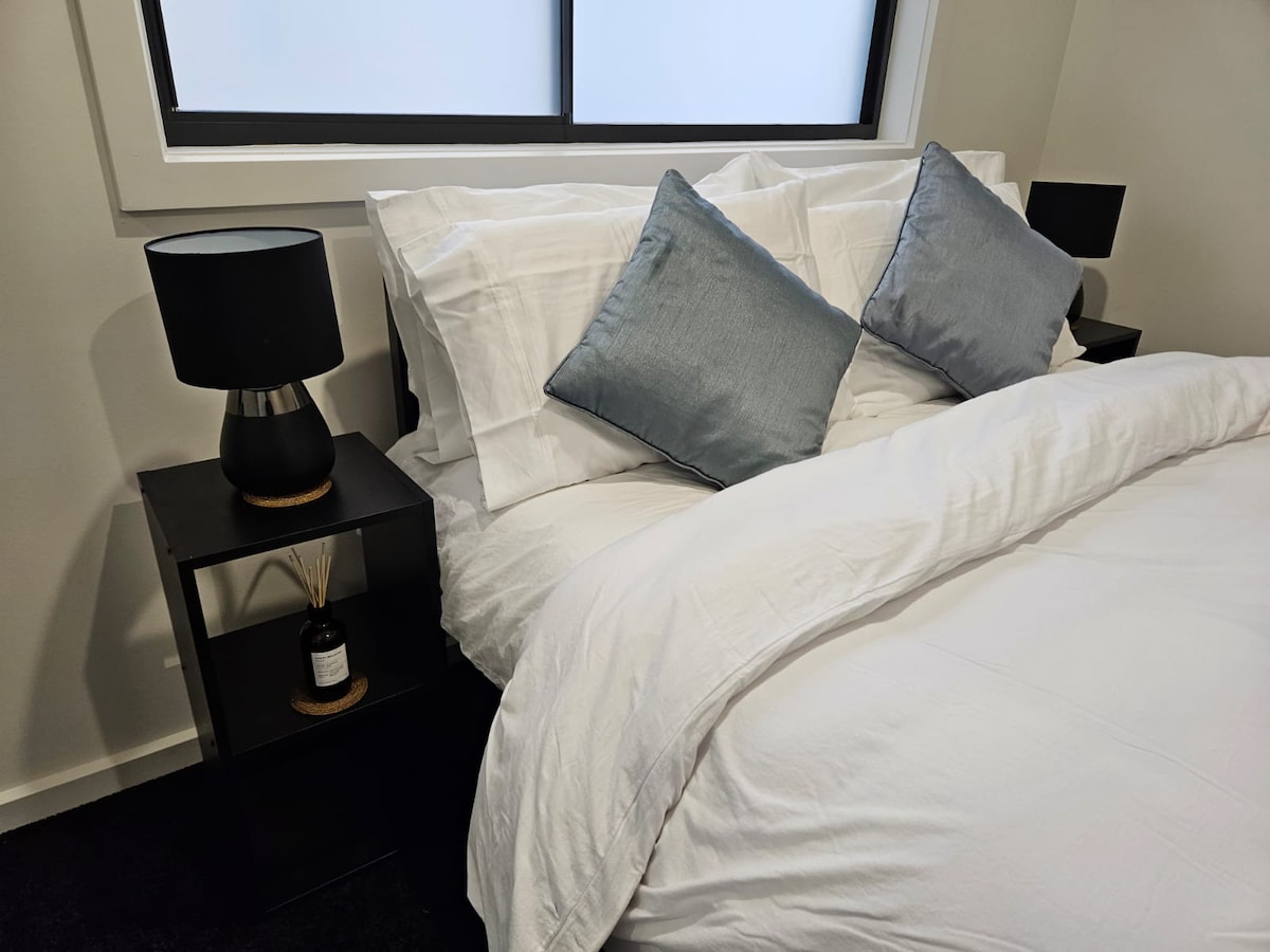 A modern bedroom features a spacious bed adorned with crisp white linens and two decorative pillows in soft blue hues. Minimalist black bedside tables hold stylish lamps, complementing the overall aesthetic. Natural light filters through a large window, enhancing the serene atmosphere.