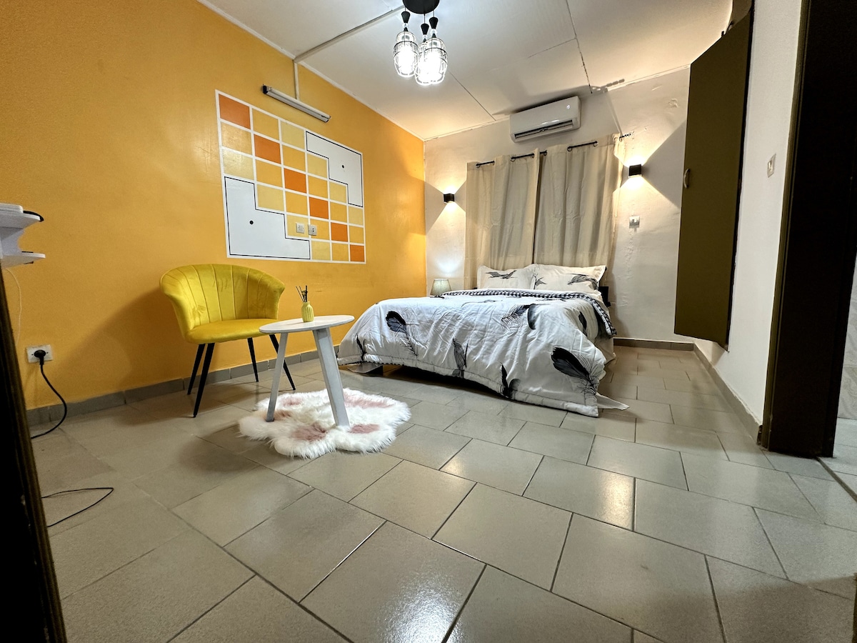 The room features a spacious bed with muted bedding, complemented by a yellow accent chair and a small white table. Light-colored walls create a bright atmosphere, while a soft rug is positioned beneath the table. Windows are dressed with neutral curtains, and climate control is visibly mounted on the wall.