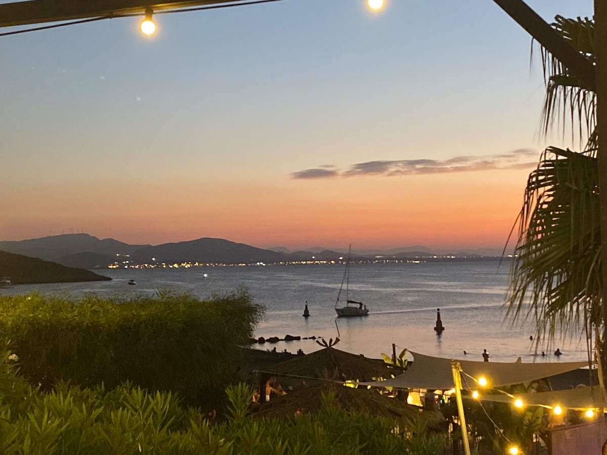 A serene view of the shoreline at sunset, featuring a calm sea with a sailboat anchored. Distant mountains are visible against a colorful sky, transitioning from orange to purple, while soft lights illuminate the foreground, creating a peaceful atmosphere.