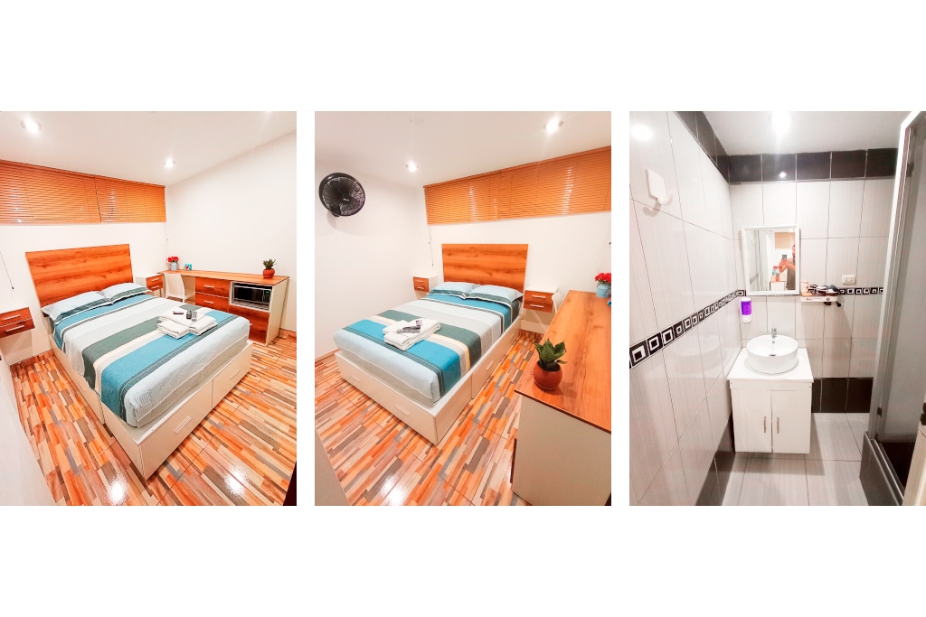 A well-furnished bedroom features a spacious bed dressed in a striped blue and white bedding set. A small plant decorates the bedside table, while a wall-mounted fan provides comfort. The room is brightly lit, highlighting its clean and simple design.