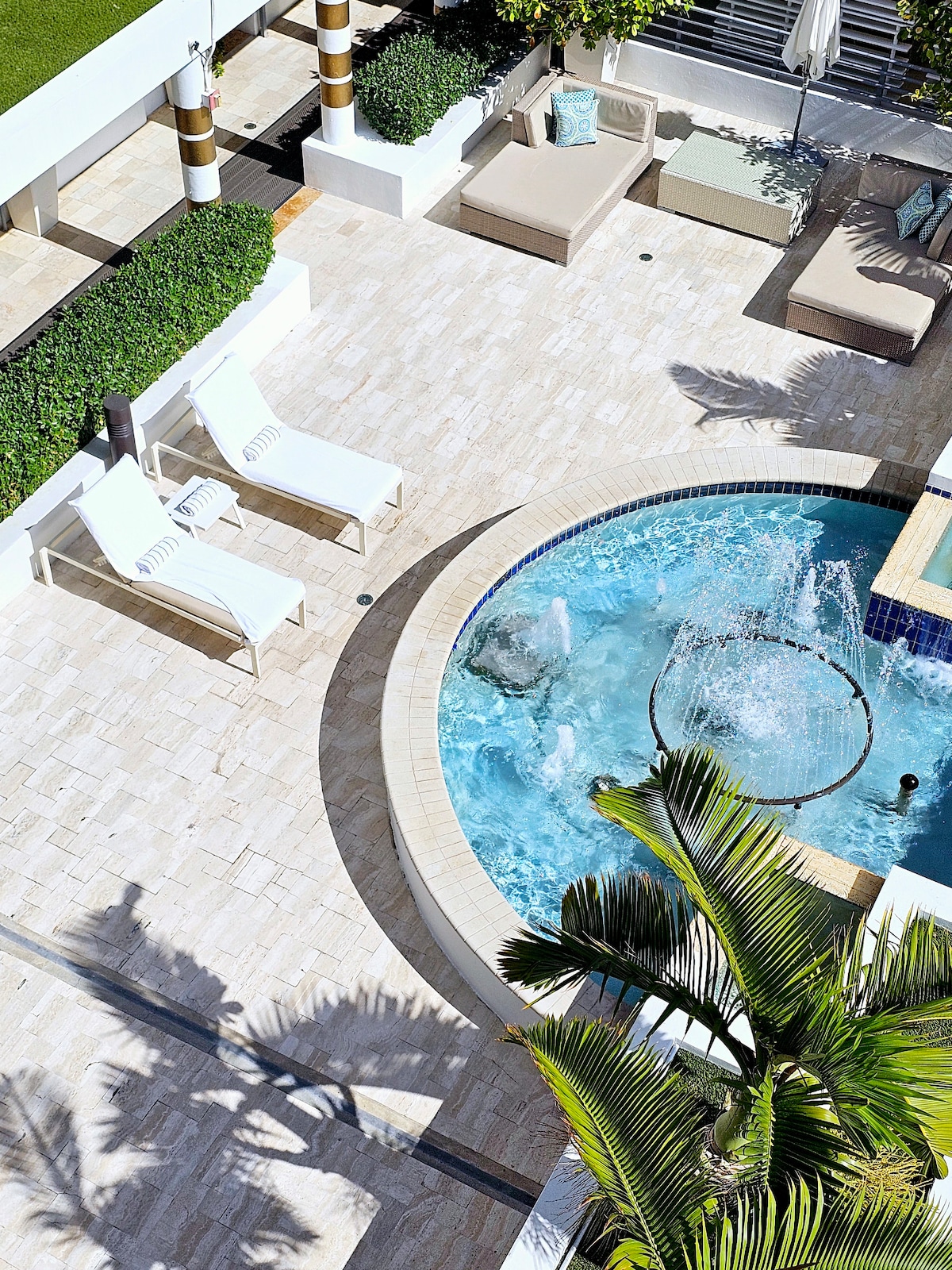 An aerial view of a shared courtyard features a circular jacuzzi surrounded by paving stones. Two white lounge chairs and a large, cushioned daybed are visible, inviting relaxation. Lush greenery and decorative columns enhance the serene atmosphere of the outdoor space.