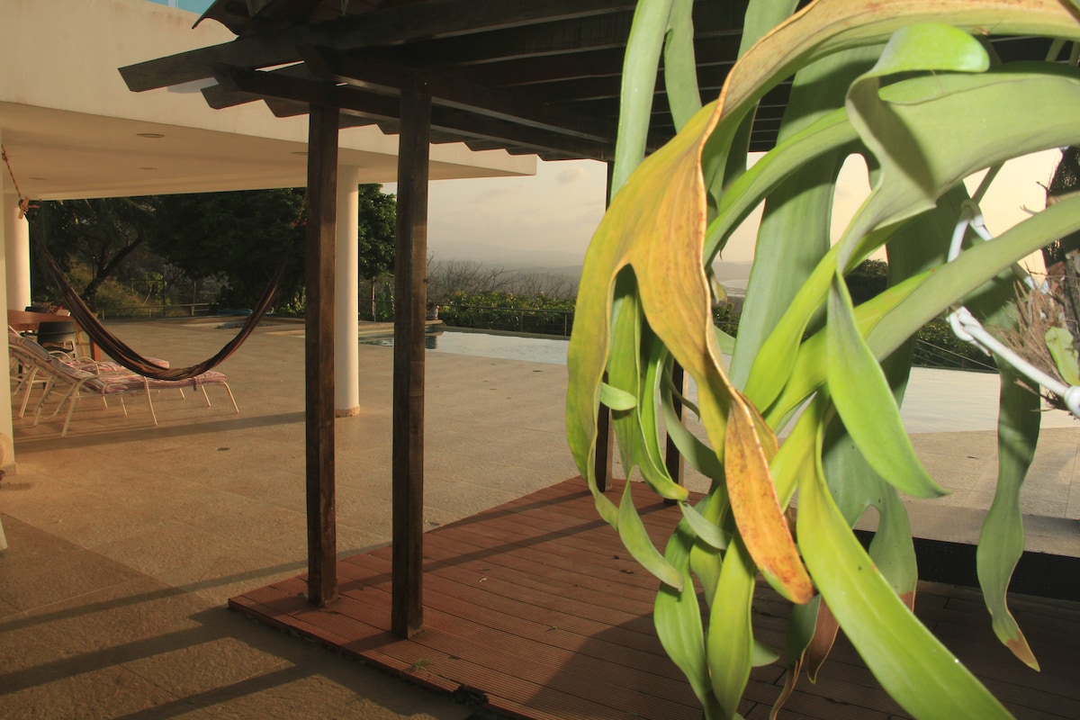 A spacious outdoor area features a shaded wooden structure with hanging plants. A hammock is visible, alongside reclining lounge chairs. The infinity pool can be seen in the background, surrounded by lush greenery and offering distant views of the Atlantic Ocean.