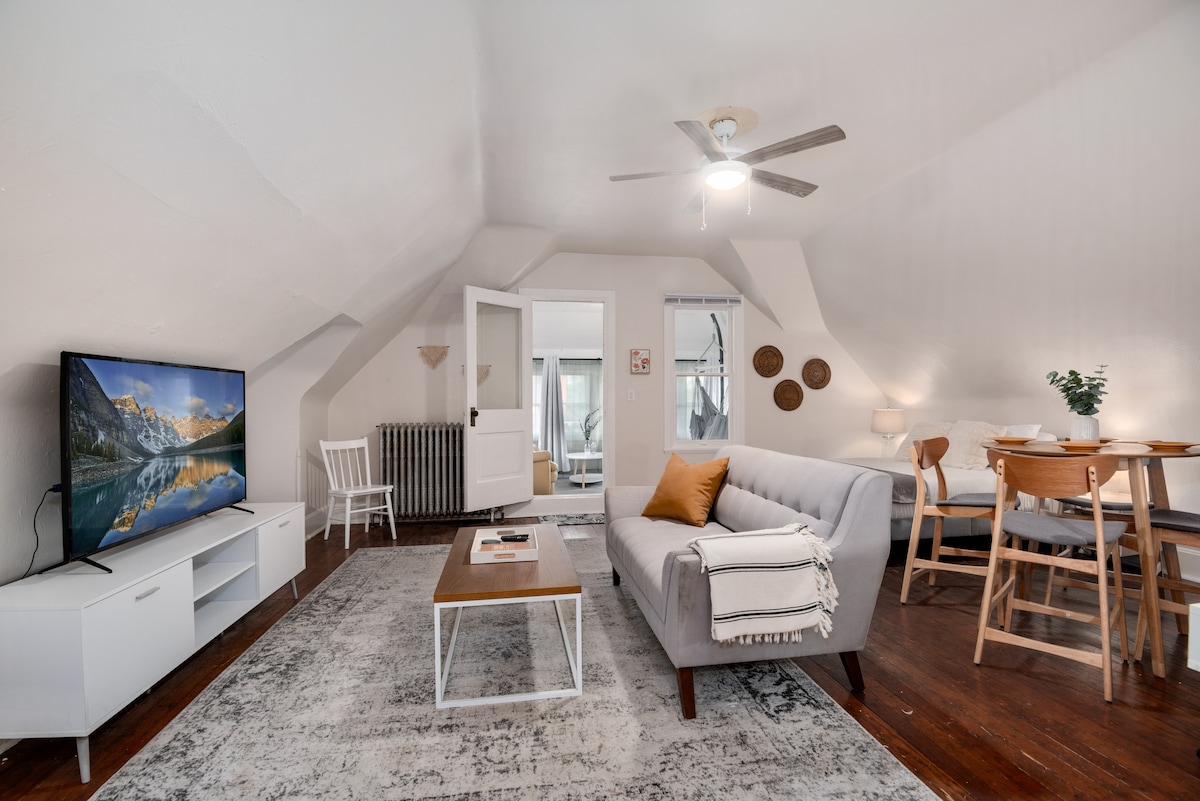 A cozy living space features a light gray sofa and a wooden coffee table placed on a textured area rug. A television is positioned on a low cabinet, and a dining area with a round table and four chairs is visible. Natural light enters through the windows.