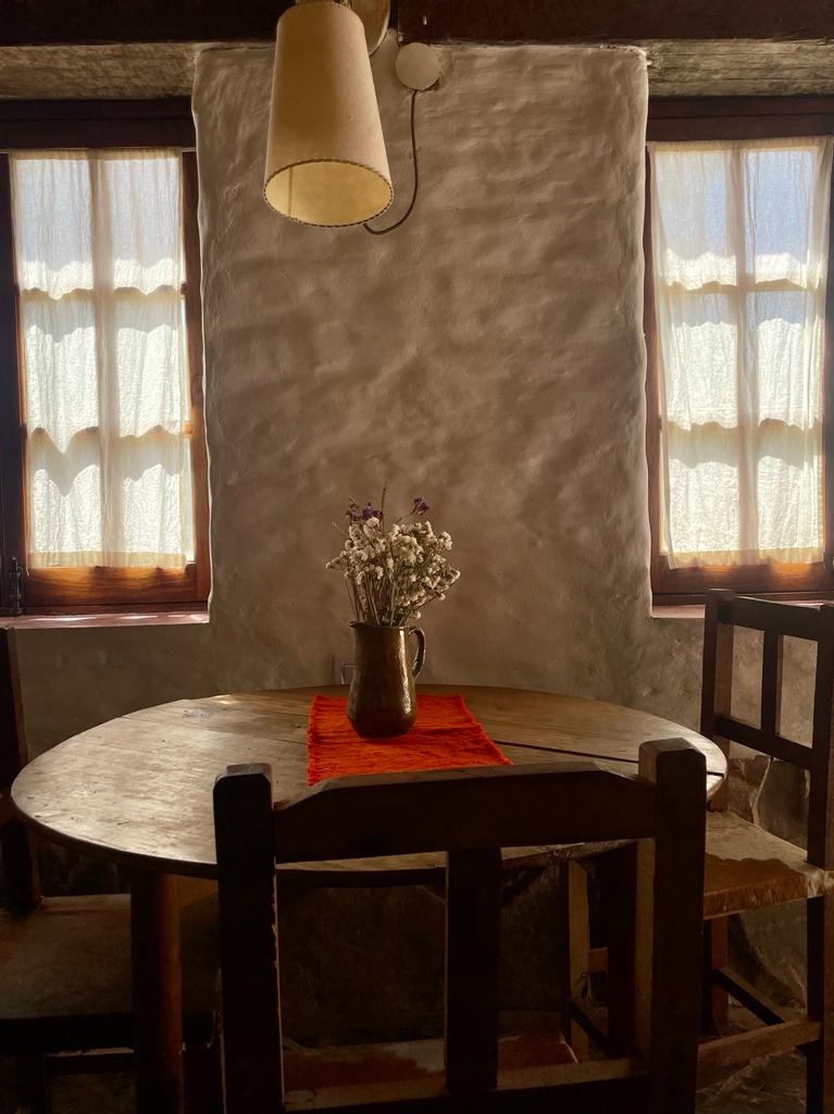 A rustic dining space features a wooden table illuminated by natural light from two windows. A simple vase with dried flowers rests at the center, complemented by a vibrant cloth underneath. The textured walls add to the cozy atmosphere of the room.