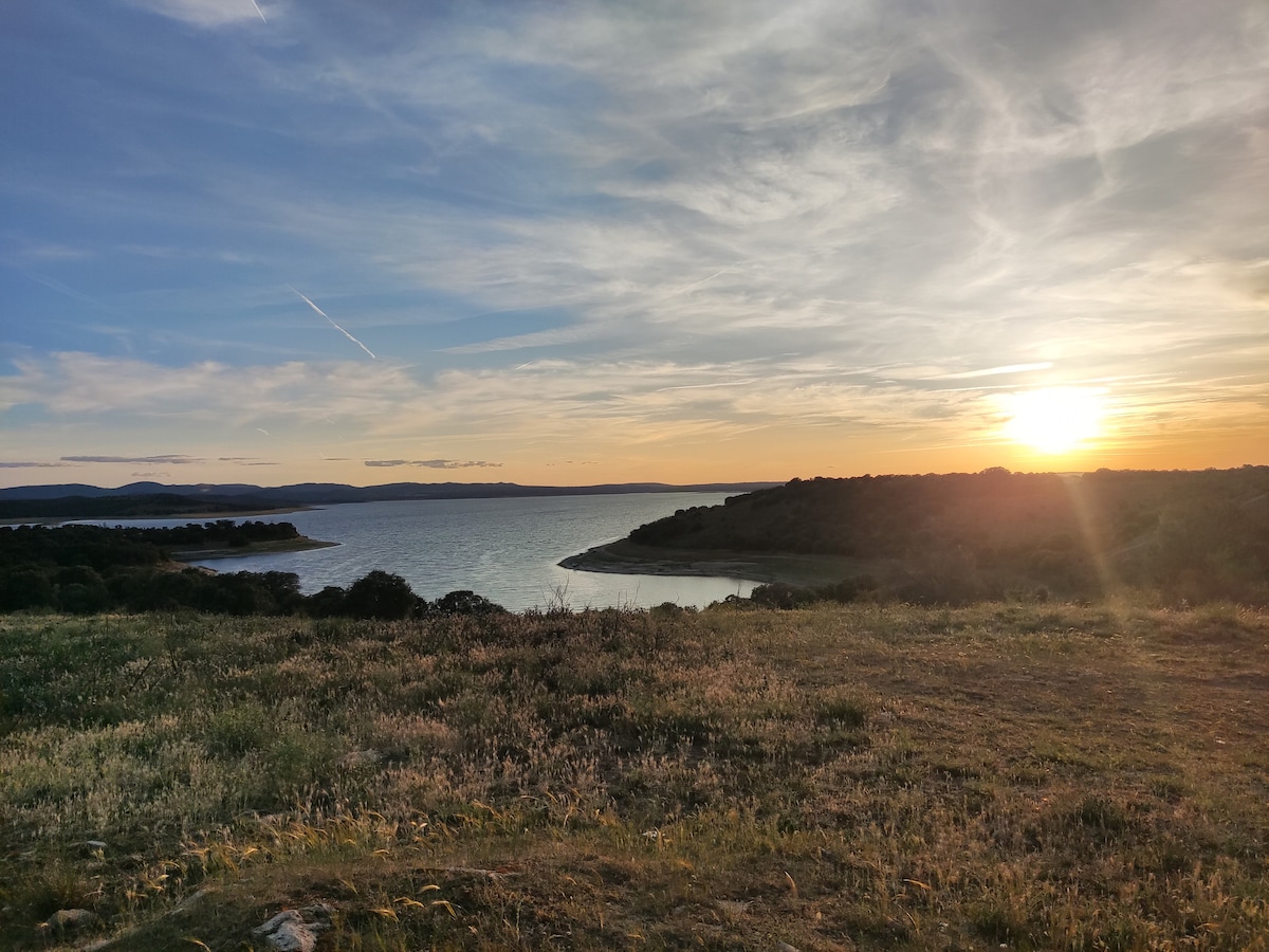 A serene landscape captures the setting sun over a calm reservoir, casting warm hues across the water. Lush grass and gentle hills frame the view, creating a tranquil atmosphere. Wispy clouds stretch across the sky, enhancing the peacefulness of the natural surroundings.