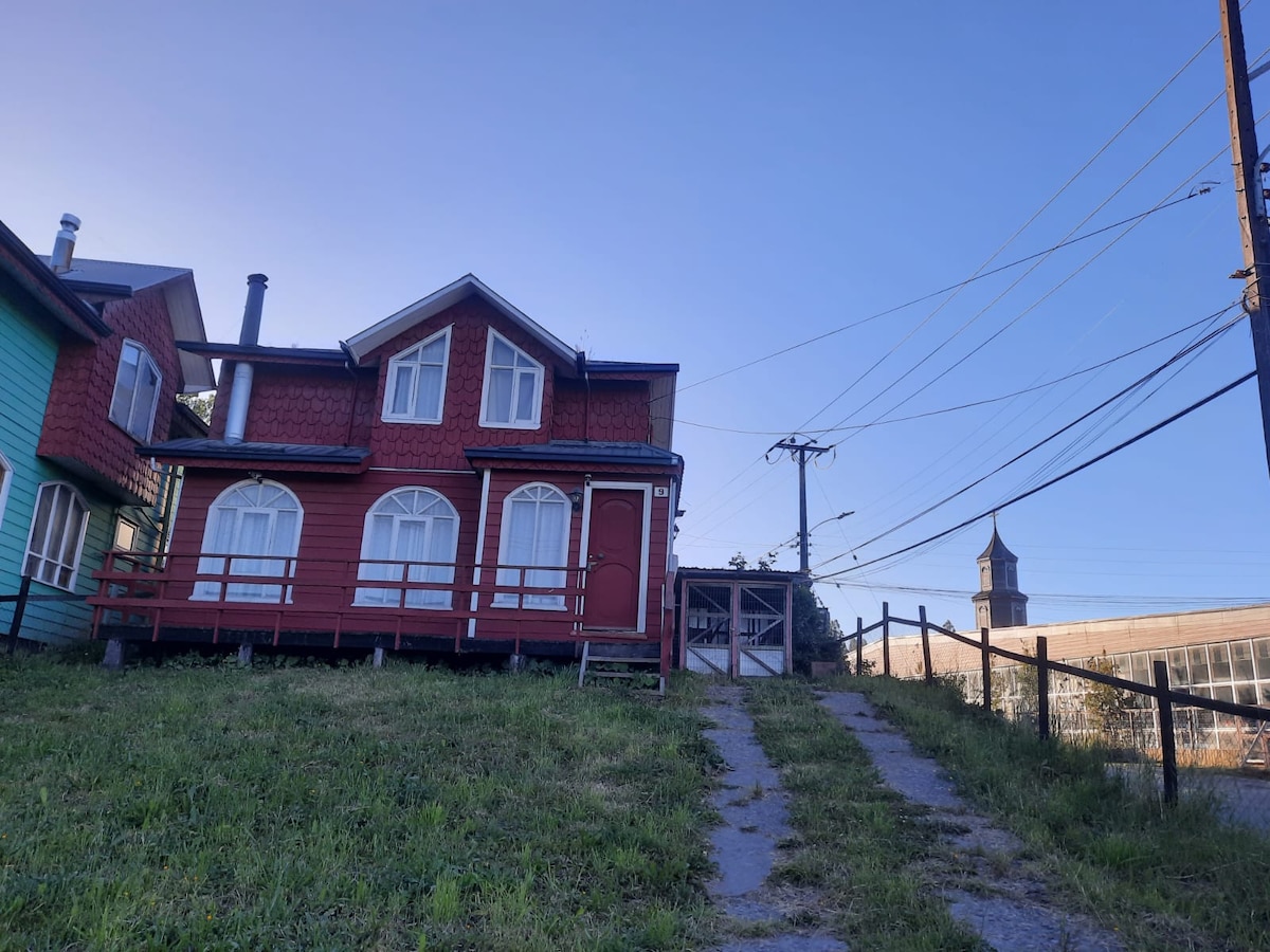 A charming red house is positioned on a grassy hill, with a sloping pathway leading to the entrance. Large arched windows are visible on the façade, and an adjacent structure, along with utility poles, can be seen in the background under a clear blue sky.
