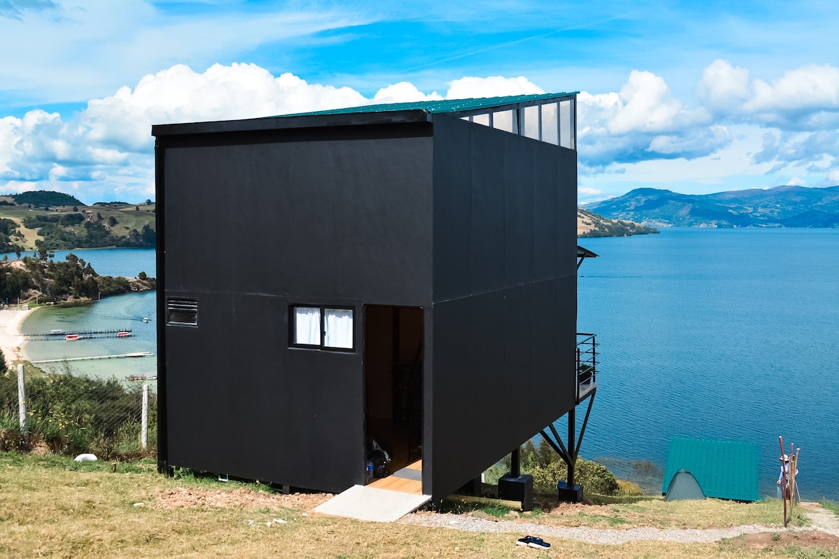 A modern black structure is positioned on a hillside, offering striking views of the lake and mountains beyond. Large glass windows illuminate the interior, while a staircase leads to the entrance. The surrounding landscape features lush greenery and a calm water's edge.