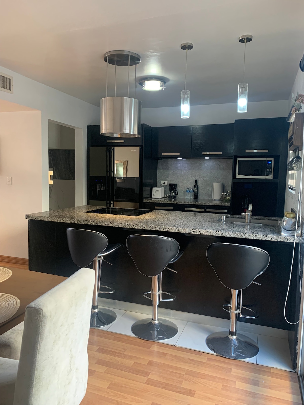 A modern kitchen features sleek black cabinetry and a spacious granite countertop. Several high-backed stools are positioned around the counter, and contemporary pendant lights hang above. Various appliances, including a microwave and coffee maker, are visible within the cabinetry.